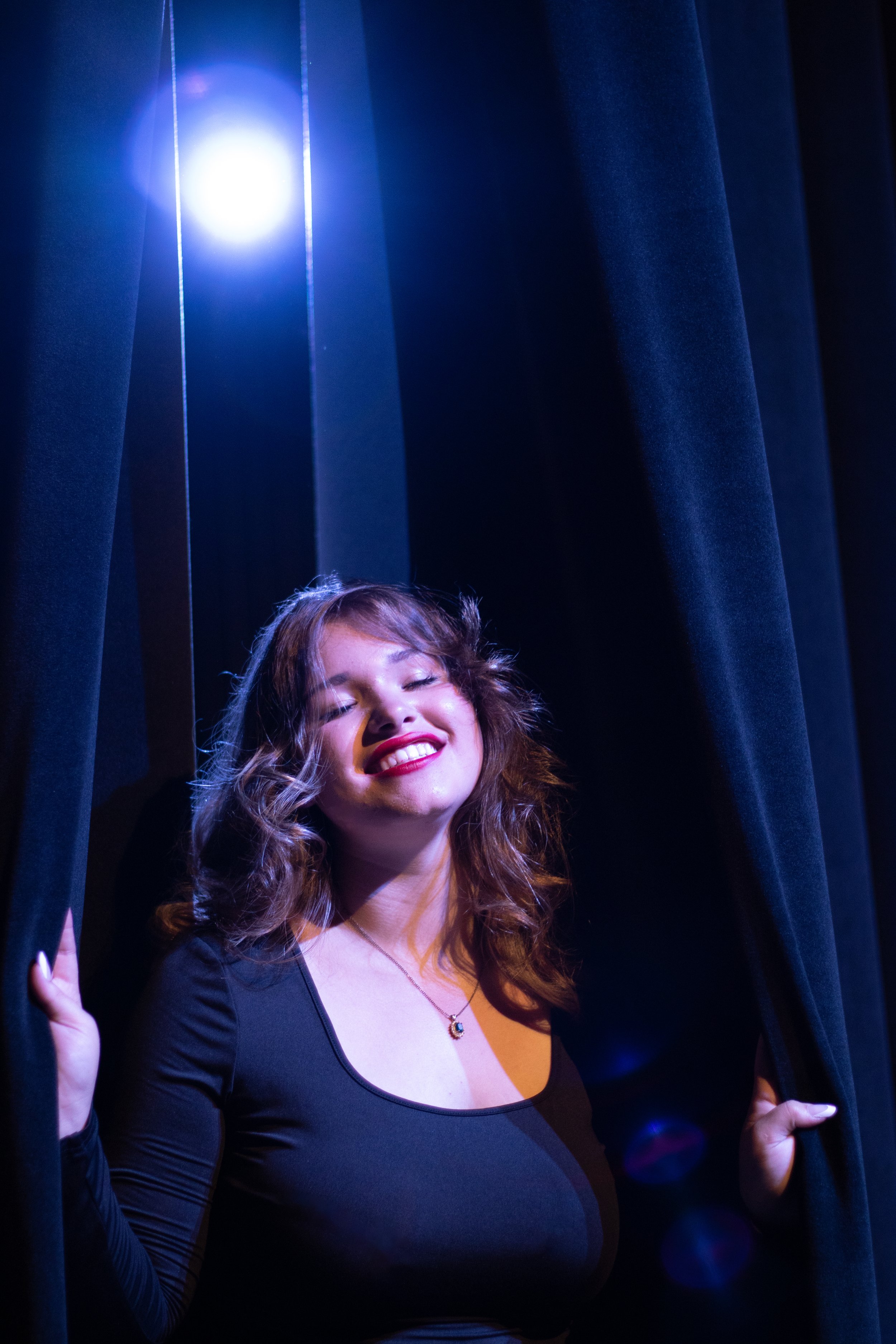 A woman with wavy brown hair and a wide smile, standing between dark blue curtains with her eyes closed, holding the curtains open as she poses on a stage with a bright spotlight shining behind her.