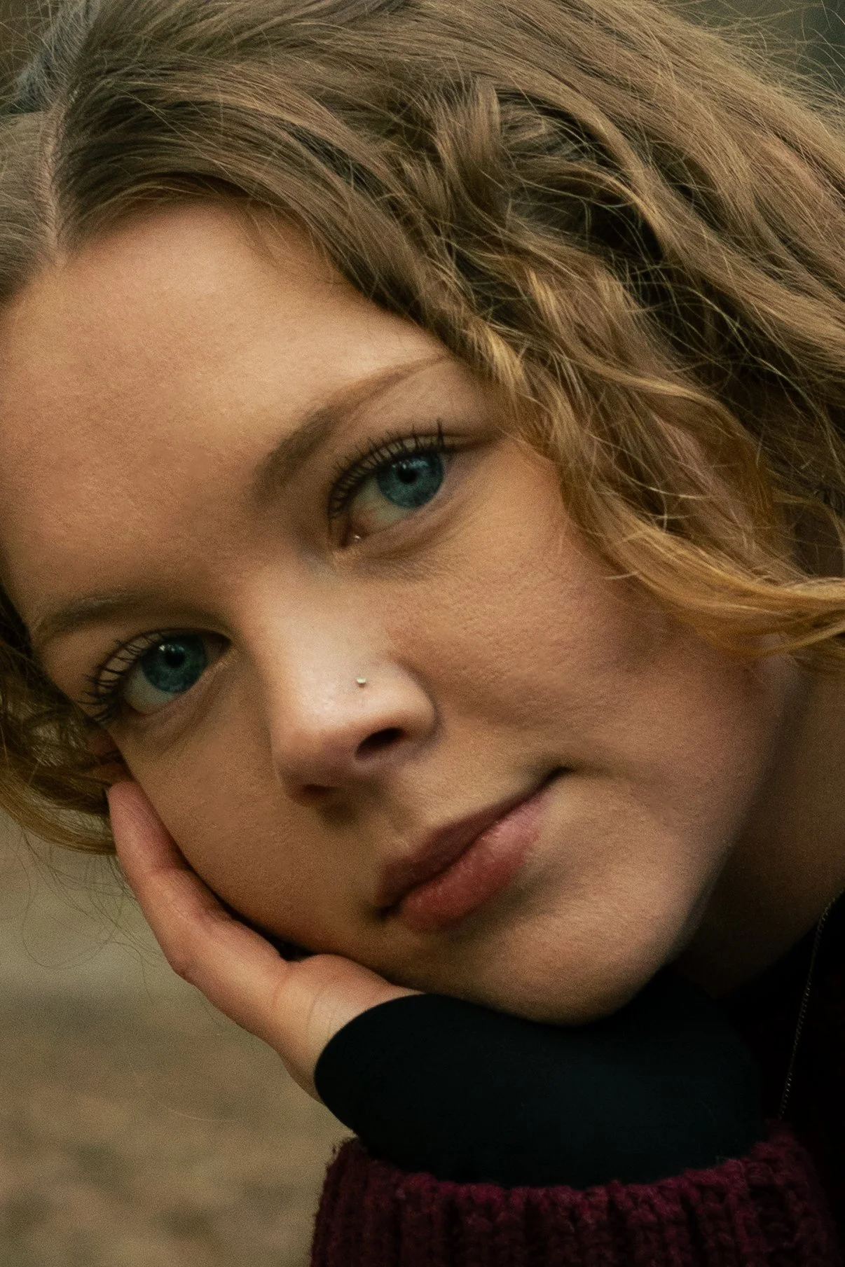 Close-up of a young woman with curly blonde hair, blue eyes, and light skin, resting her face on her hand, wearing a dark top and a maroon sweater.