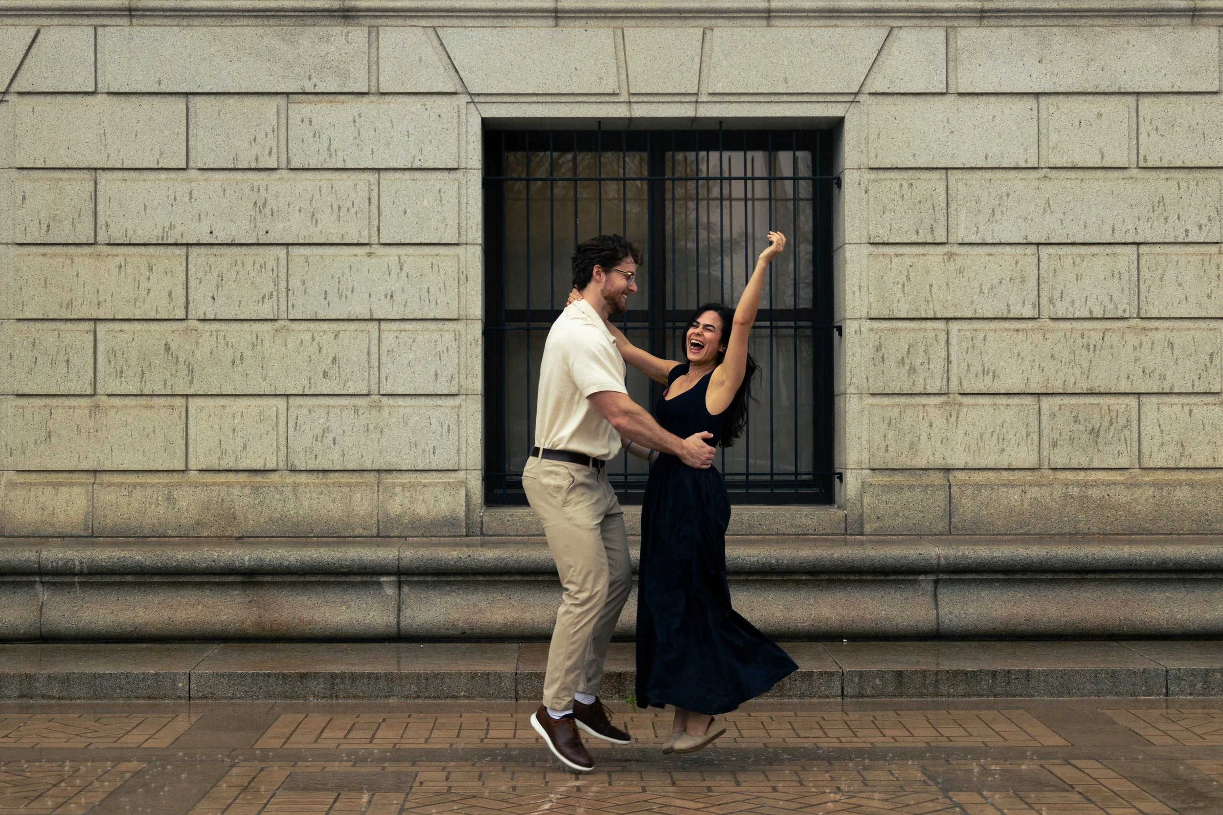 A man and woman joyfully dancing and hugging on a sidewalk in front of a beige brick wall with a barred window.