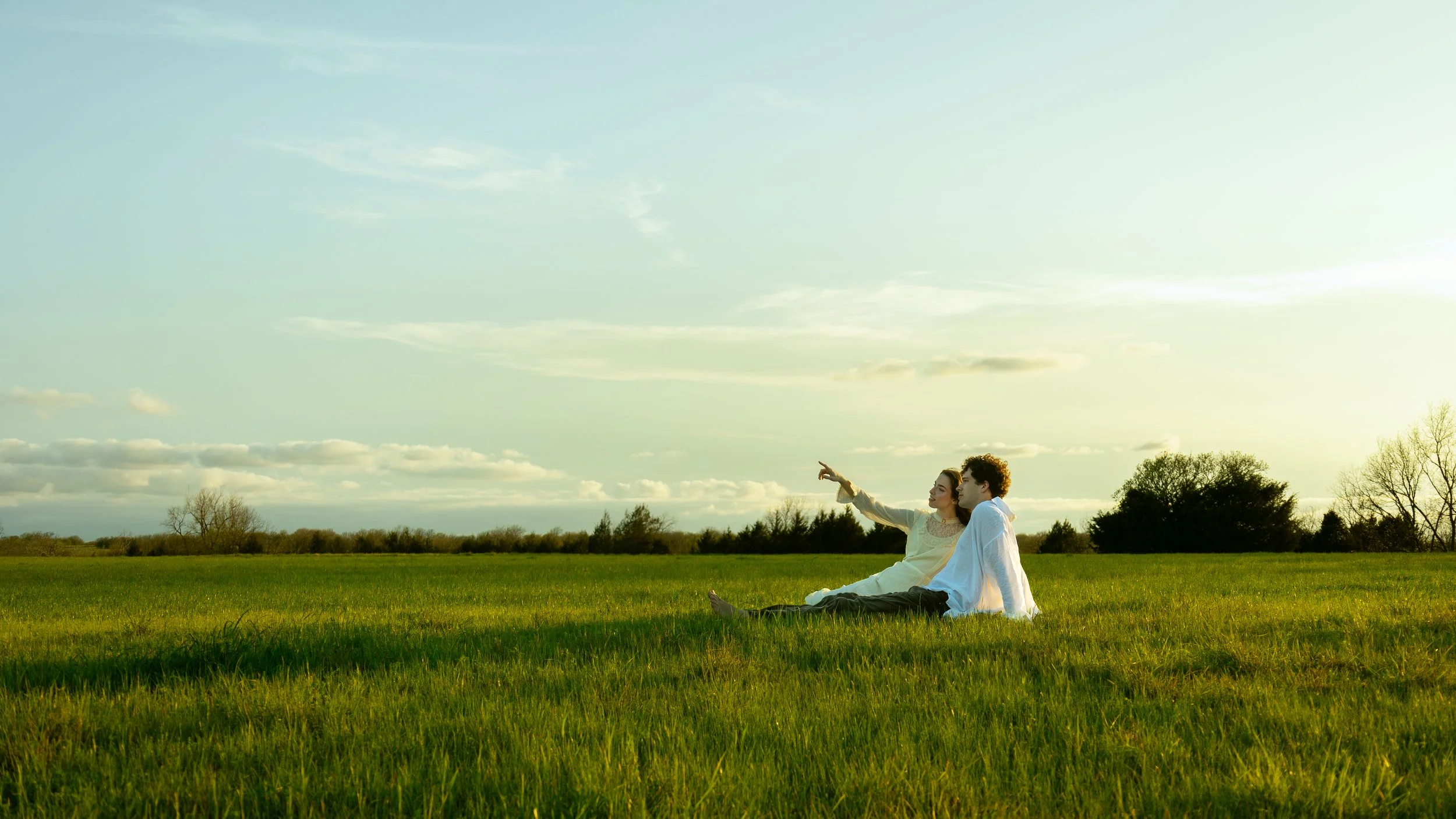 A couple sitting on a grassy field, with the woman pointing towards the sky during sunset or late afternoon.
