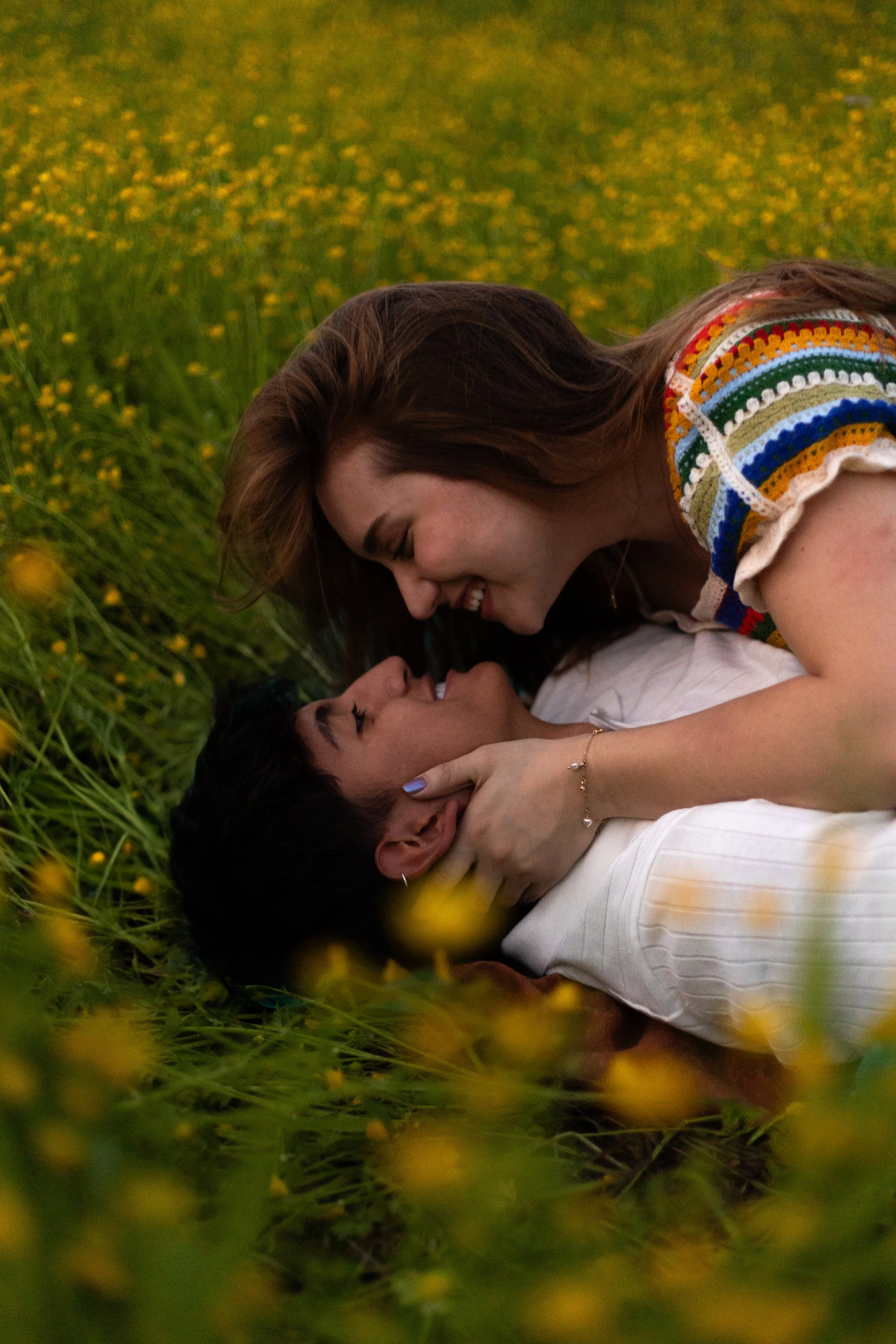 A couple lying on the grass in a field of yellow flowers, smiling and leaning close to each other.