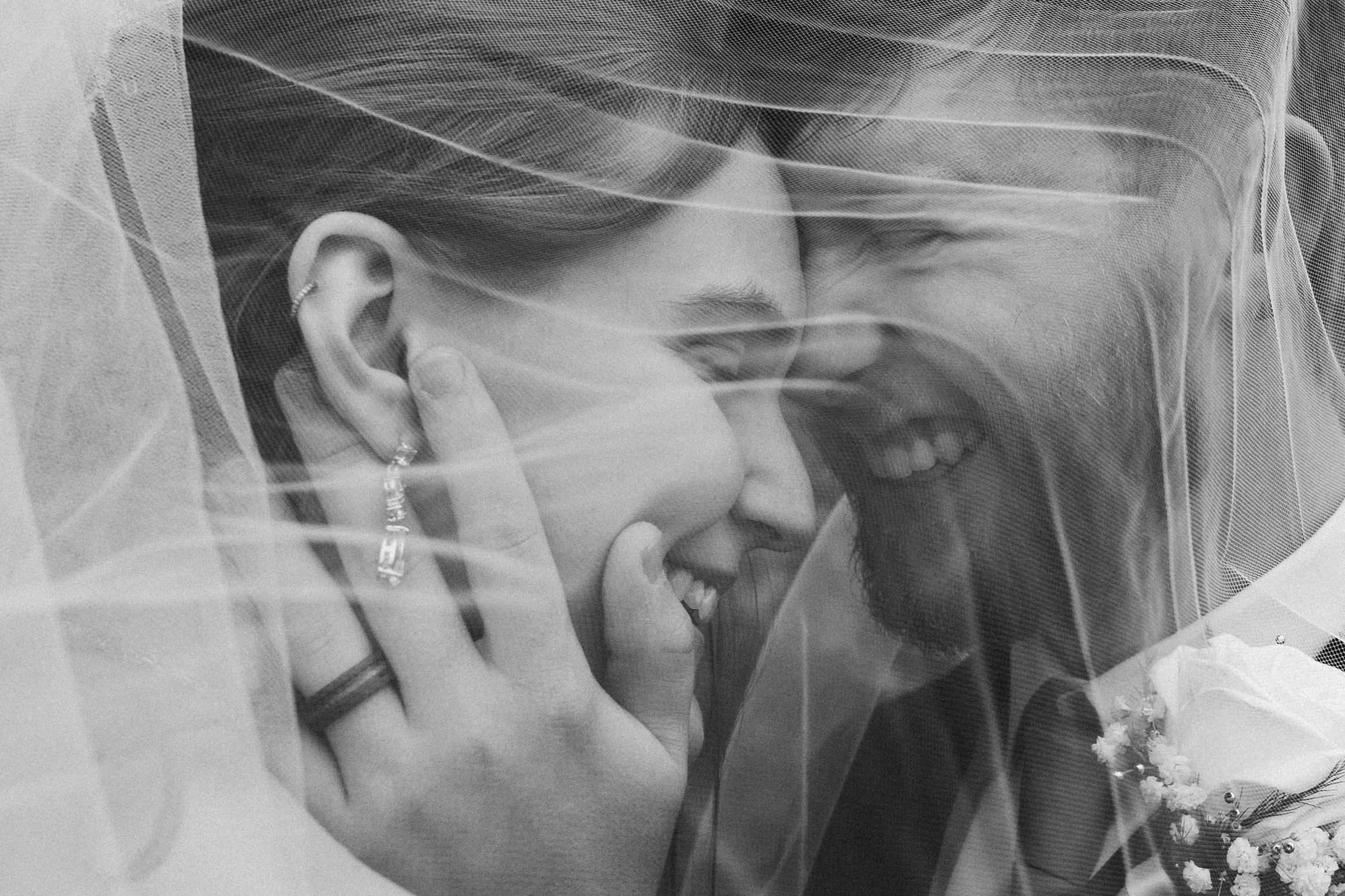A black and white photograph of a smiling woman and a man behind a wedding veil. The woman's face is close to the man's, and she is touching her ear with her hand, showing earrings and a ring. There is a white flower boutonniere on the man's lapel.
