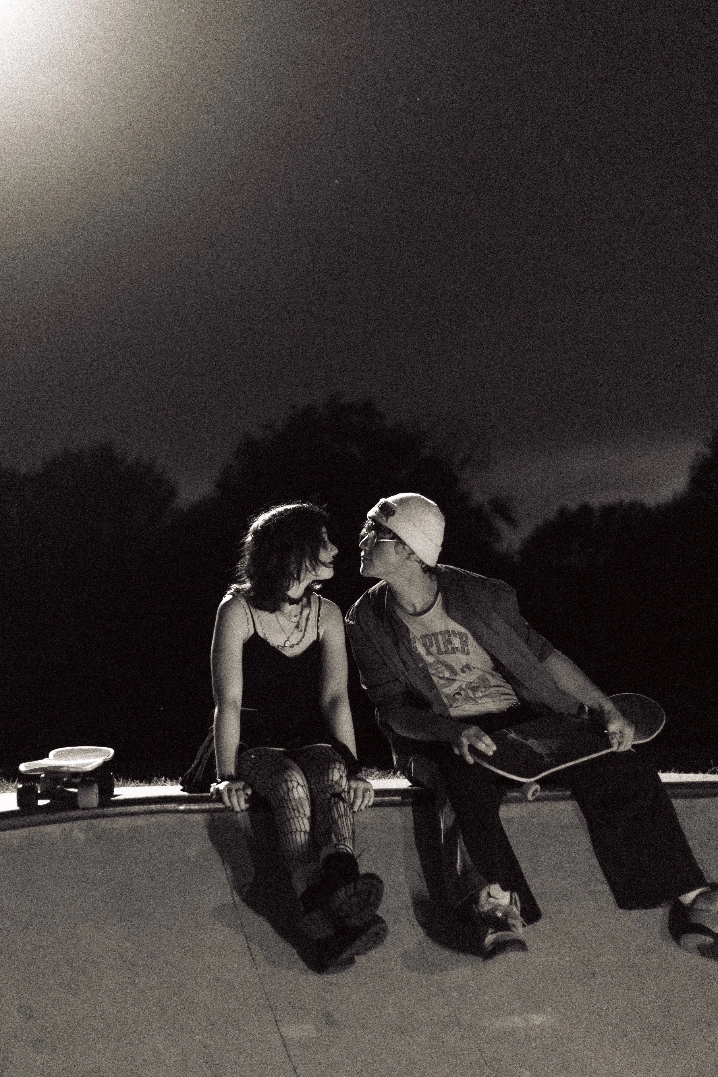 A black and white photo of a young couple sitting close together on the edge of a skatepark rim at night, with the woman wearing a sleeveless top and fishnet stockings, and the man wearing a beanie, glasses, and a jacket, holding a skateboard.
