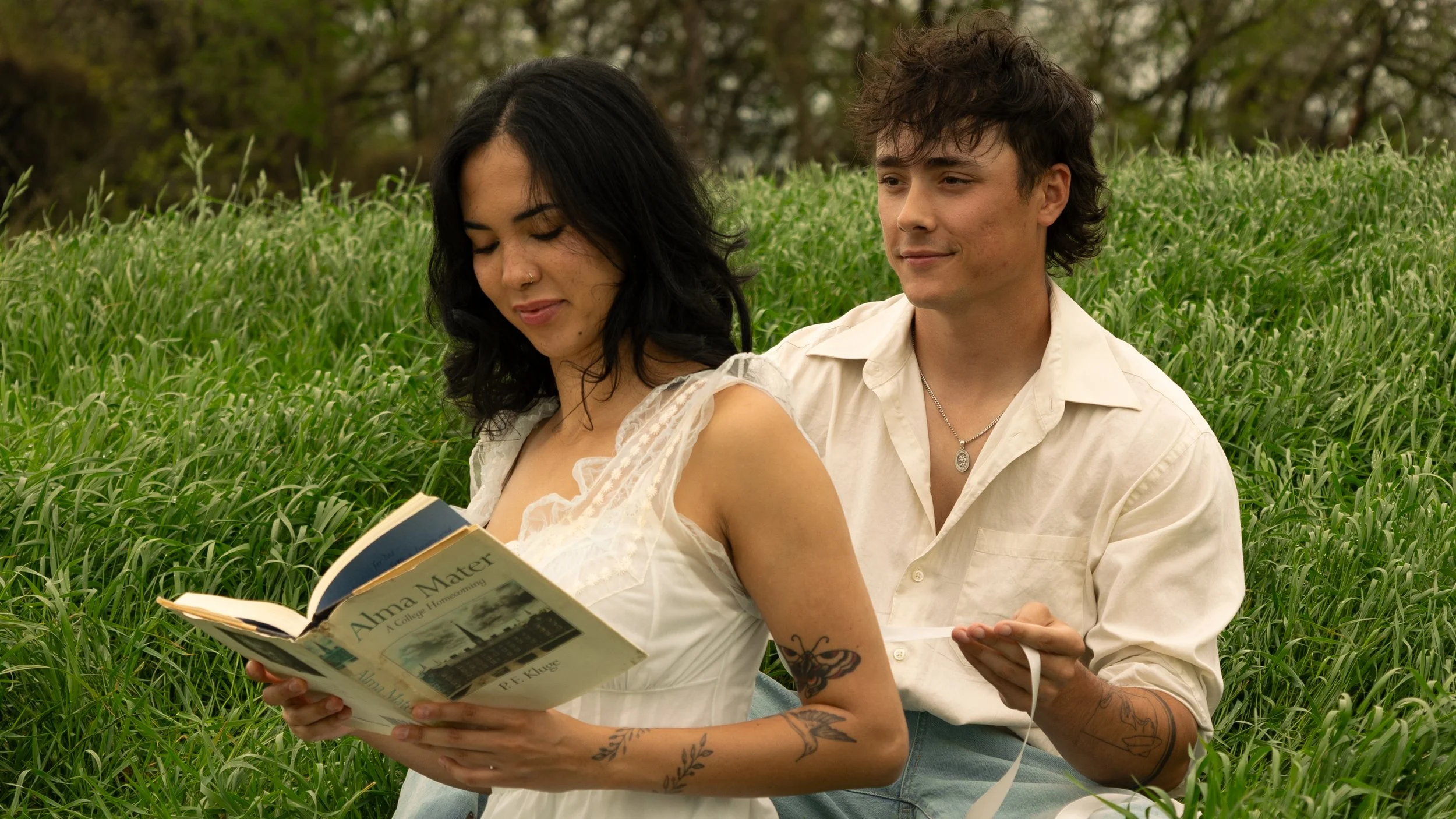 A woman with black hair, tattoos on her arm, and wearing a white dress, sitting on grass and reading a book titled 'Alma Mater' by P. F. Kluge. A man with curly hair, tattoos on his arm, and wearing a cream shirt, sits behind her holding a white ribb