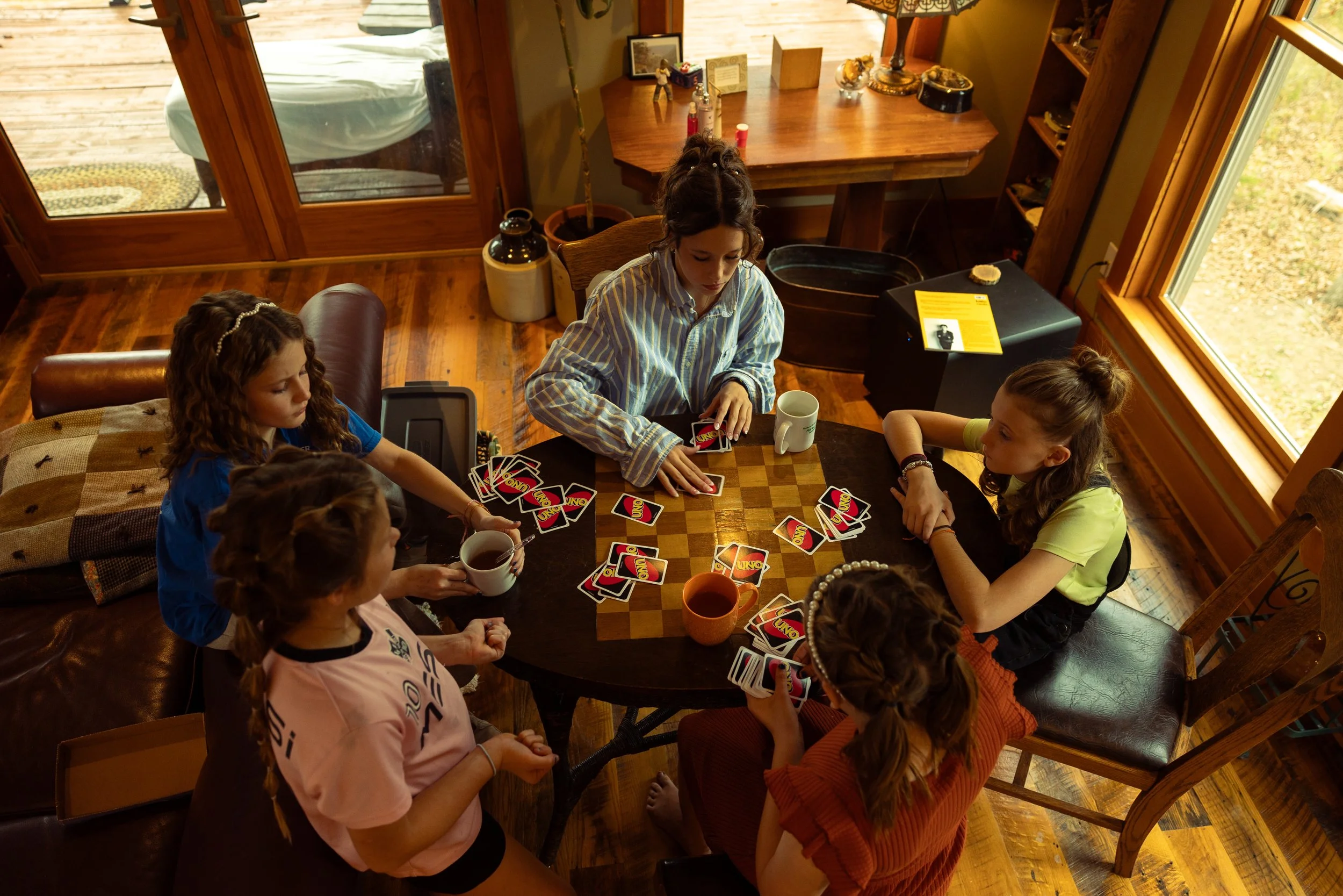 Six children sitting around a wooden table playing the card game UNO with two cups and a mug of tea, in a cozy room with wooden floors, large windows, and shelves with decorative items.
