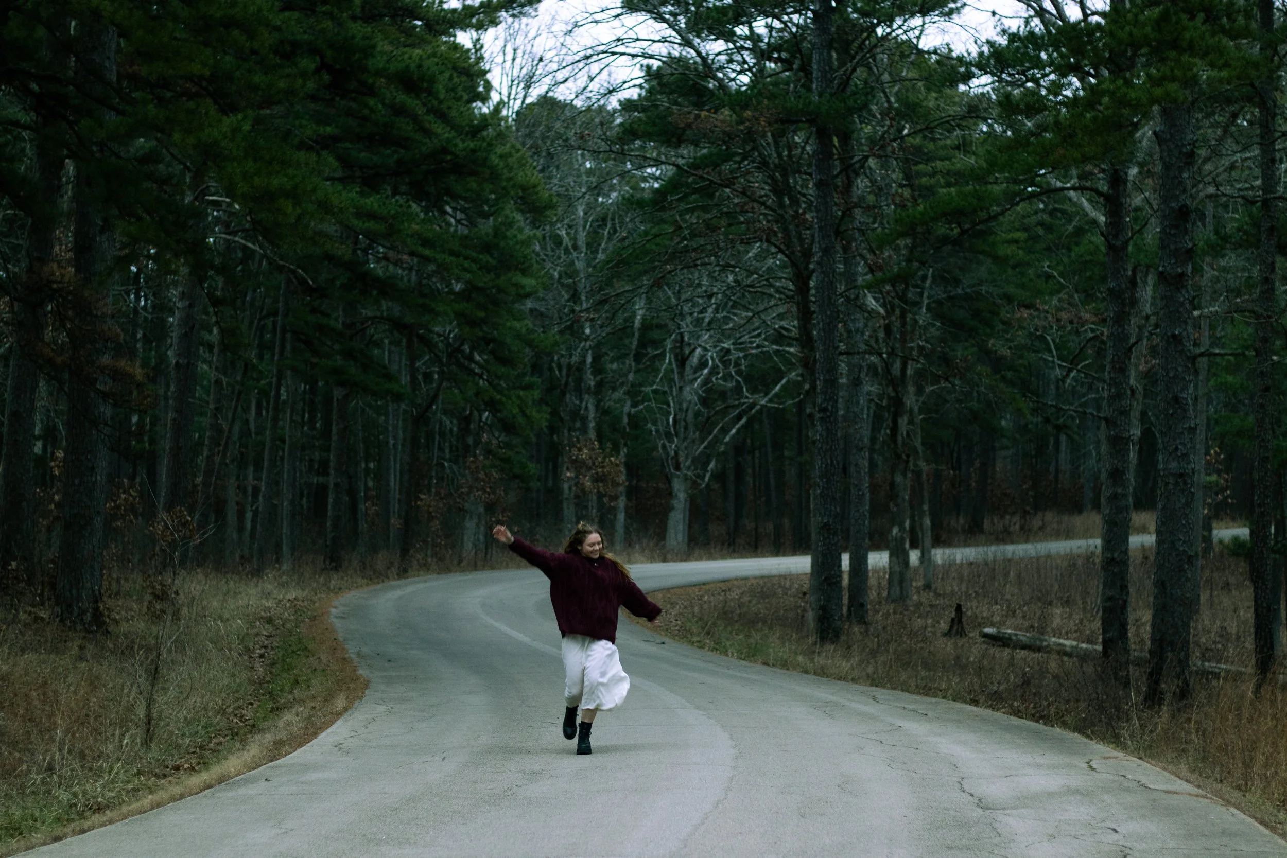 A woman walking and smiling on a winding forest road surrounded by trees in autumn or late fall.