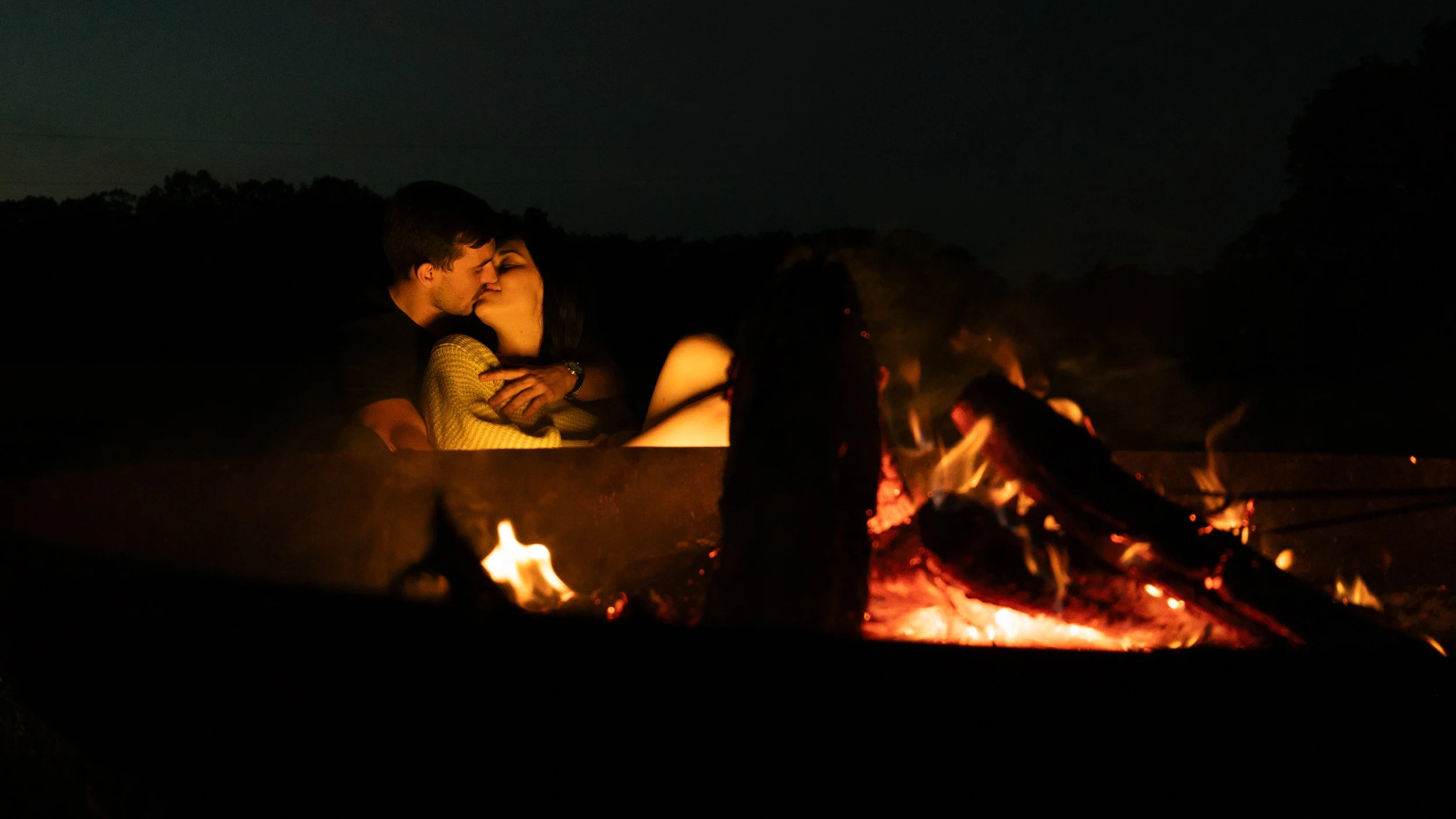 A couple is sitting close together outside at night, sharing a kiss near a campfire with glowing embers, trees, and a dark sky in the background.