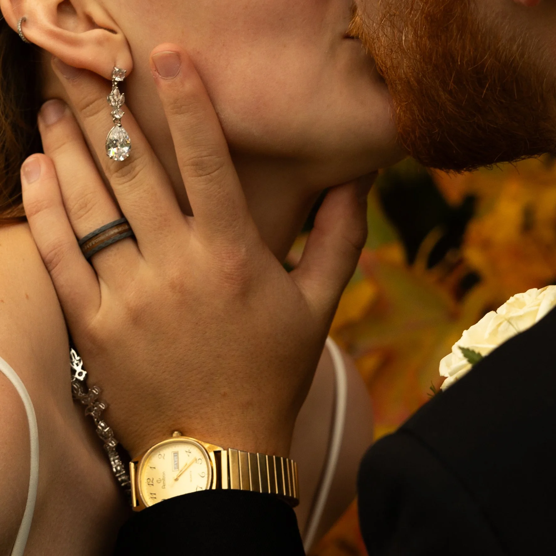 Close-up of a couple kissing, showing the woman's hand with rings, an earring, a bracelet, and a gold watch, and the man in a suit.
