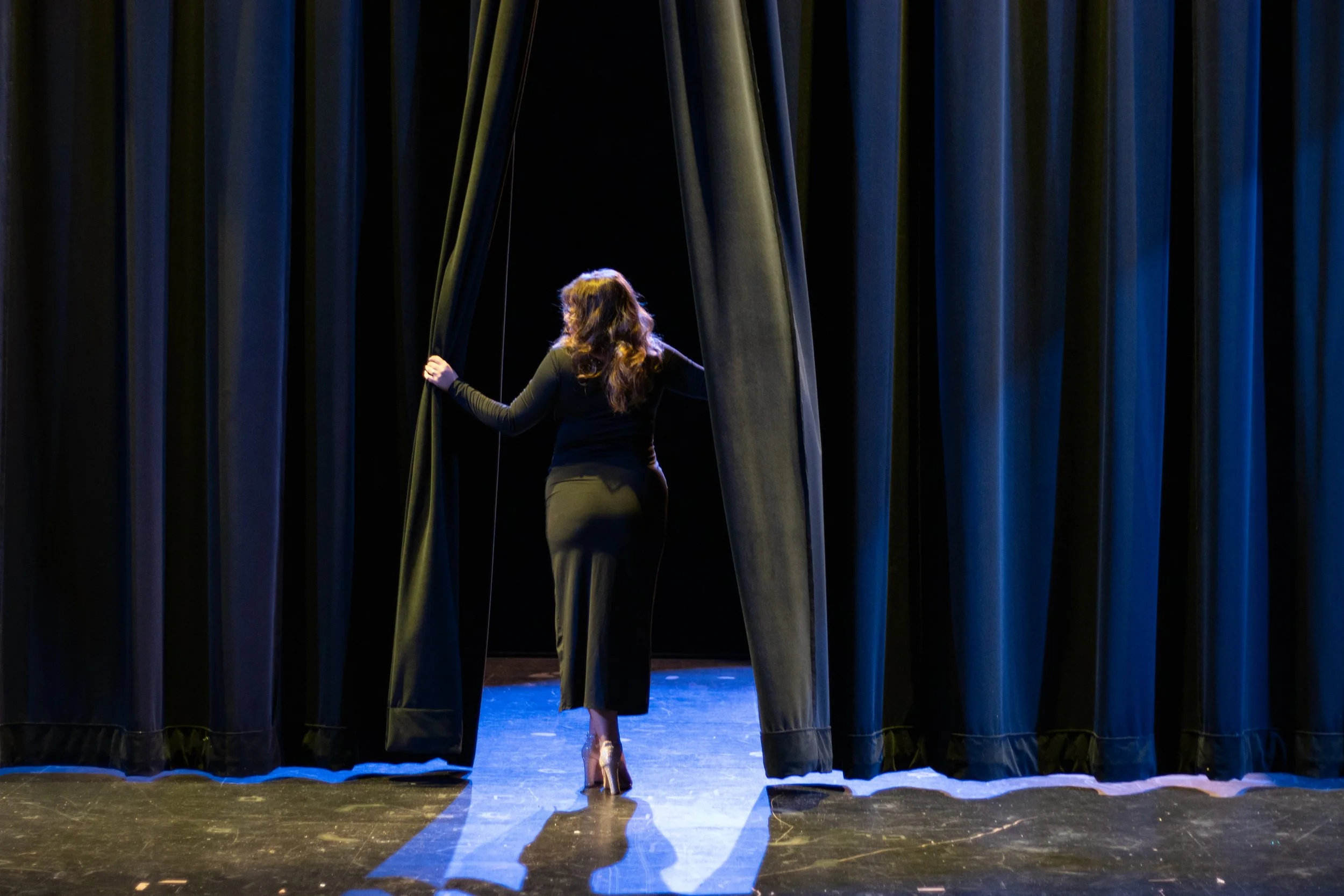 A woman with long hair, dressed in black, walks onto a stage through black curtains, with their back facing the camera and holding the curtains open.