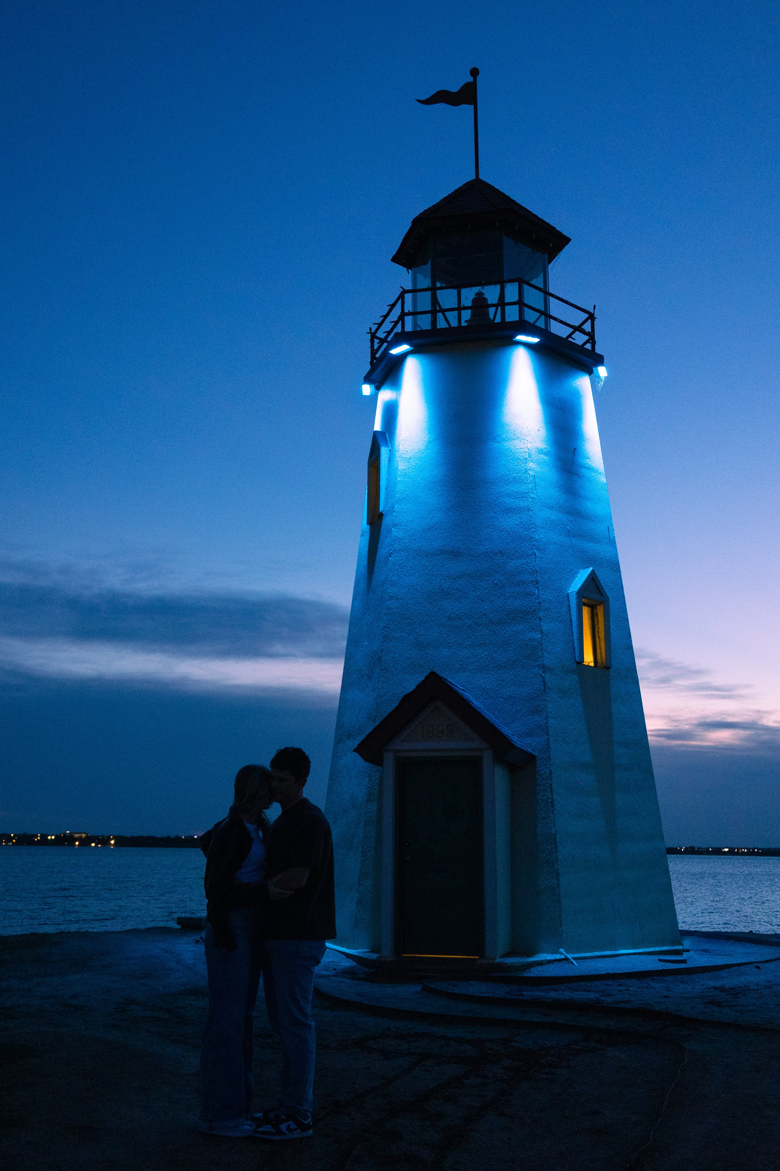 Couple standing close together near a lighthouse illuminated with blue lights at dusk by the water.