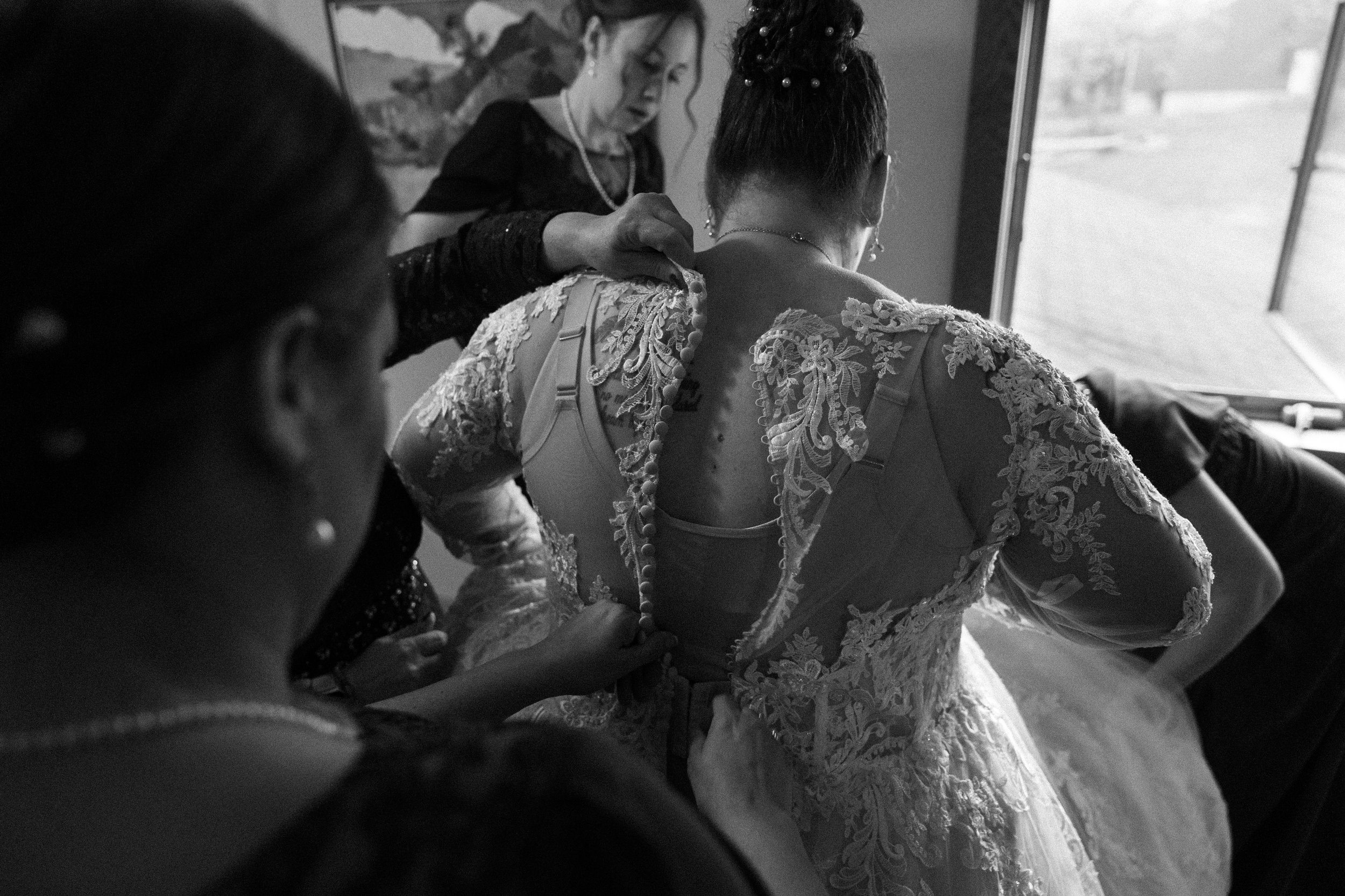 Woman in a lace wedding dress being assisted by others as she prepares for her wedding, seen from behind, near a window.