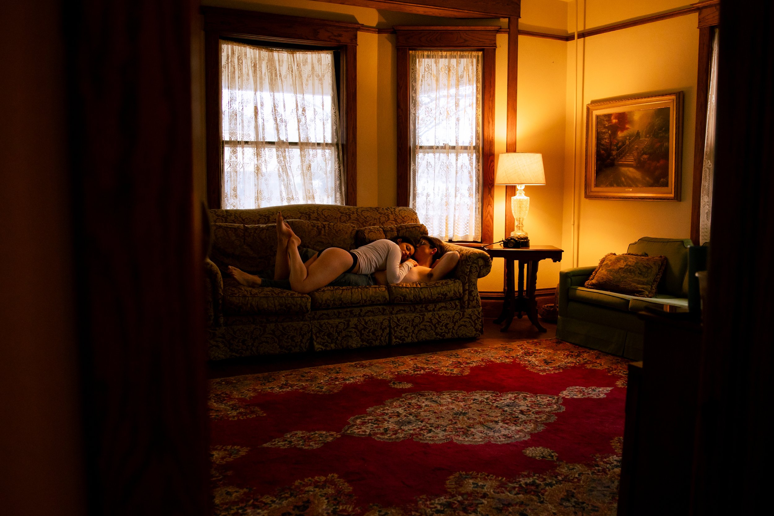 A woman and a man lying on a patterned sofa in a cozy, warmly lit living room, engaged in an intimate moment. The room features curtains over windows, a side table with a lamp, a framed painting, and a green armchair on a red floral carpet.