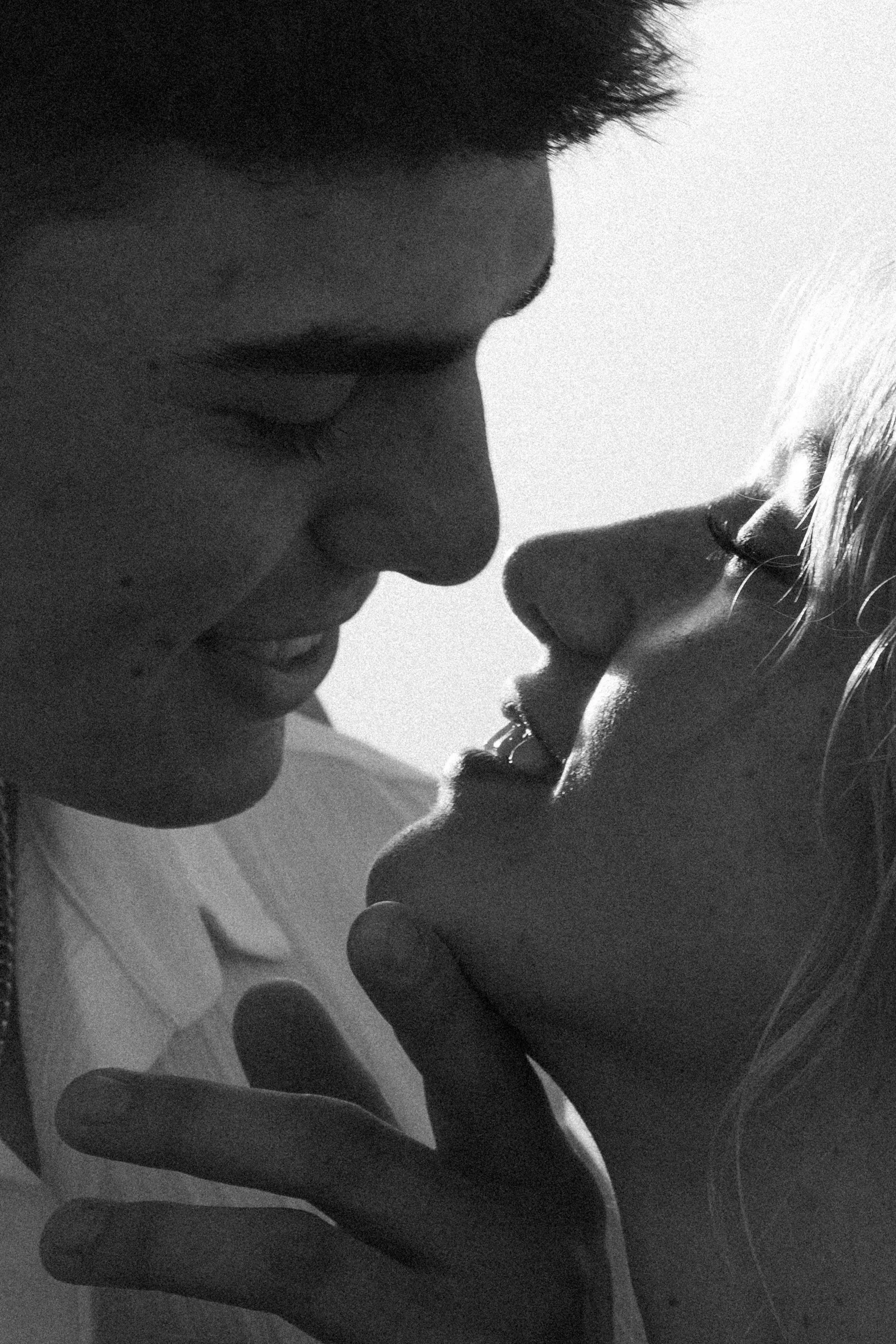 A black and white close-up photograph of a couple about to kiss, with their faces very close, showing their profiles and with a soft background.