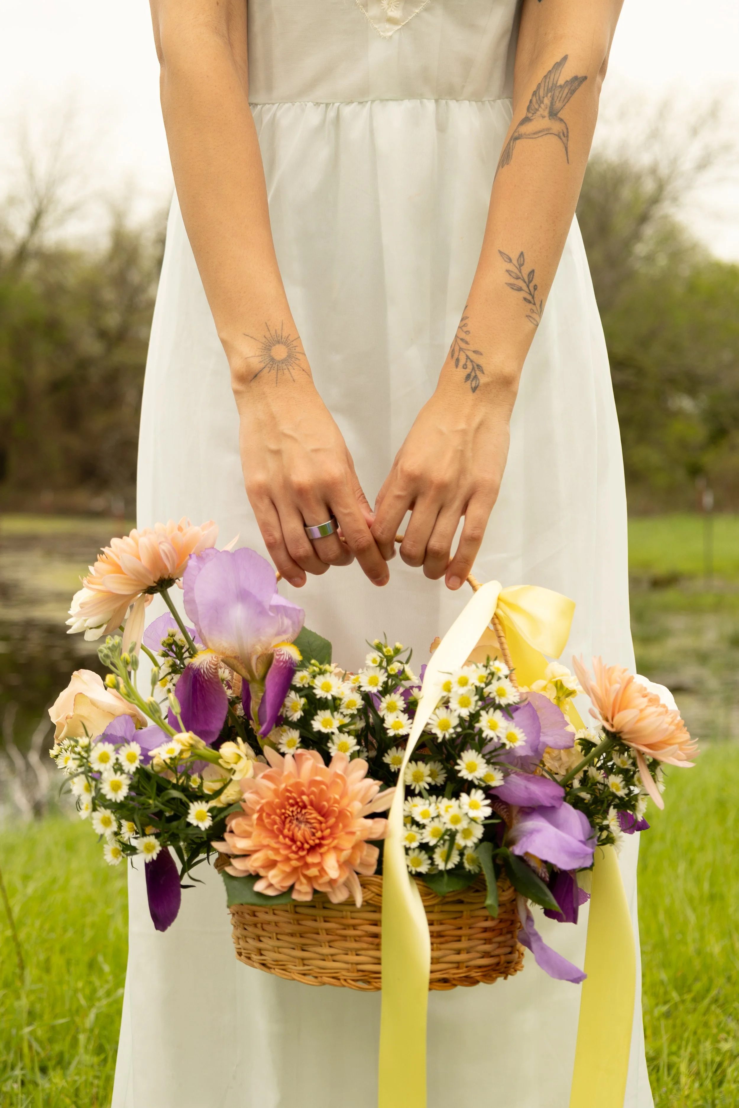 A woman in a white dress holding a basket of colorful flowers outdoors, with tattoos on her arms.