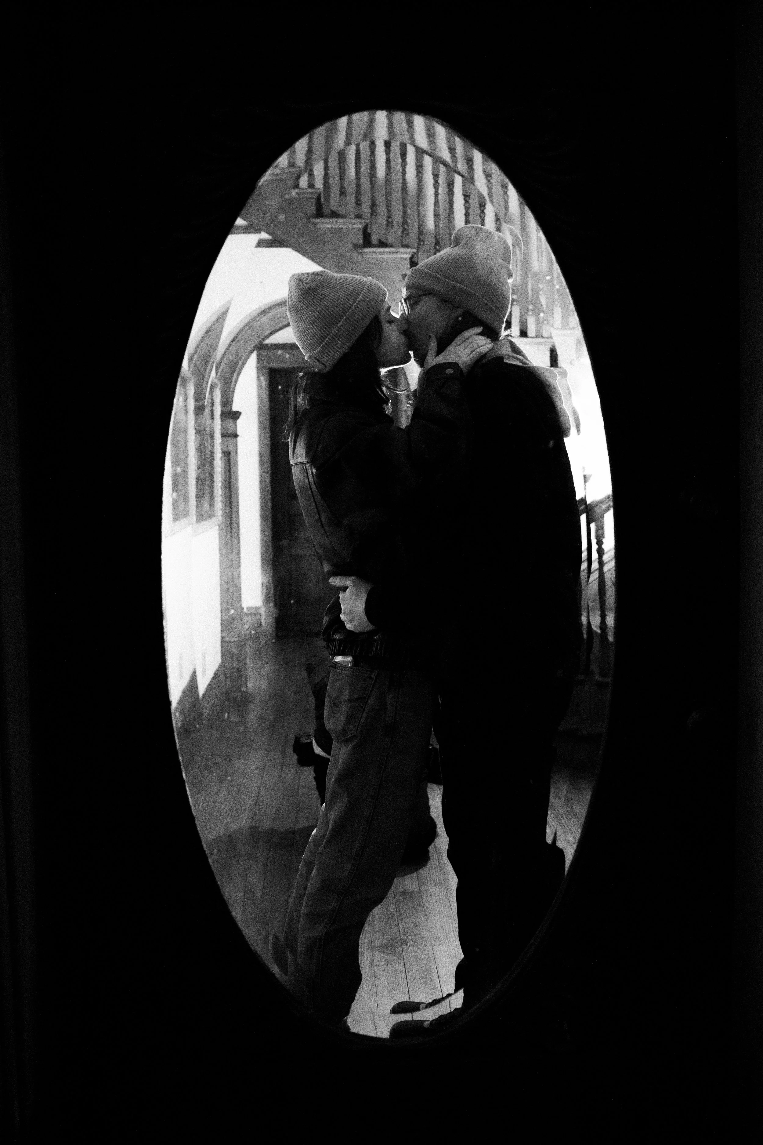 A black-and-white photo of a couple kissing, viewed through an oval-shaped mirror. They are wearing beanies and winter clothing inside a cozy home with wooden floors and a staircase in the background.