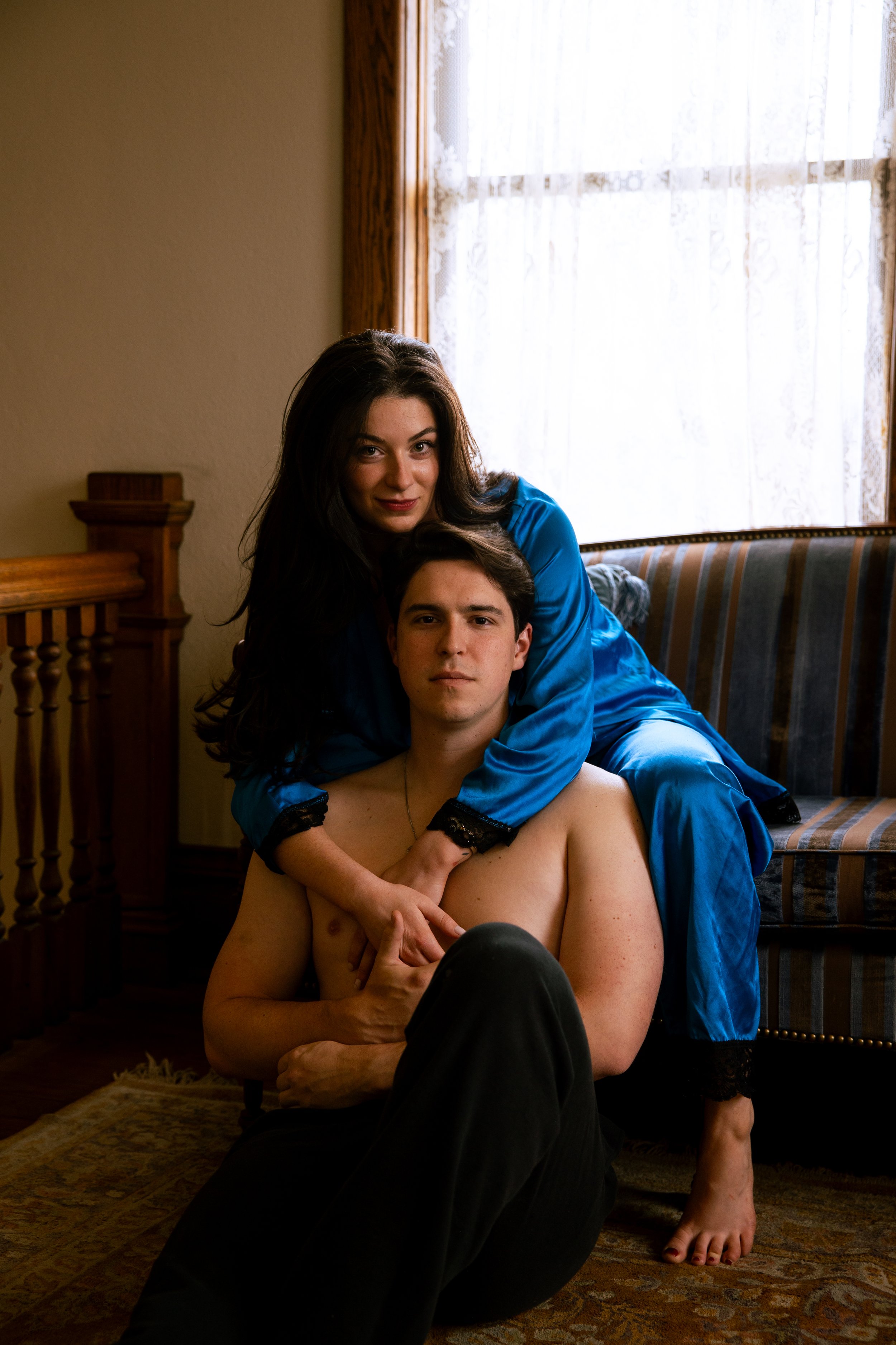 A woman in a blue satin dress sitting on a couch, embracing a shirtless man with dark hair, black pants, and a necklace, in a room with a wooden floor, beige walls, and a window with lace curtains.