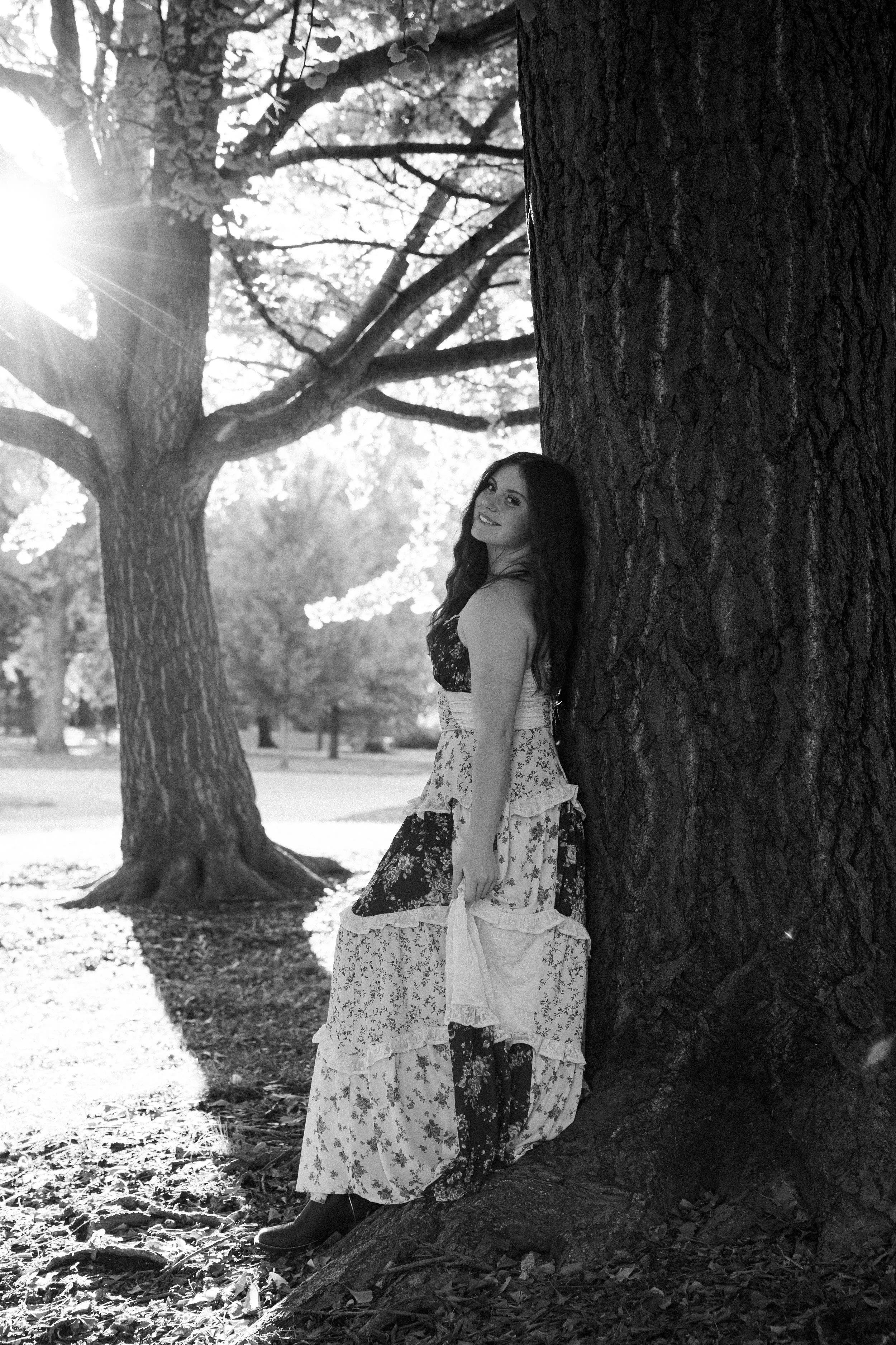 A woman in a floral dress leaning against a tree in a park, with sunlight streaming through the branches.