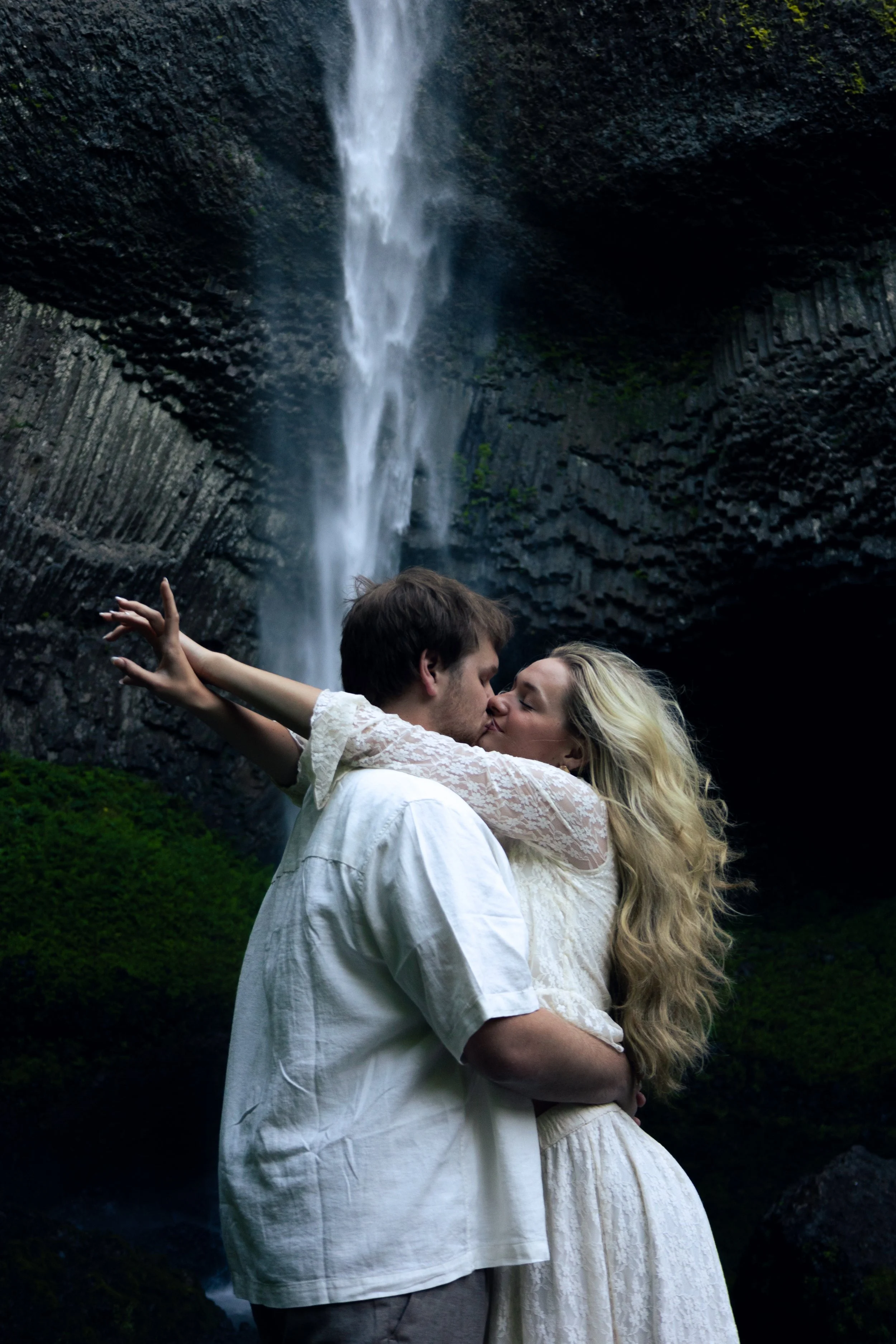A couple kissing in front of a waterfall surrounded by dark rocks and greenery.