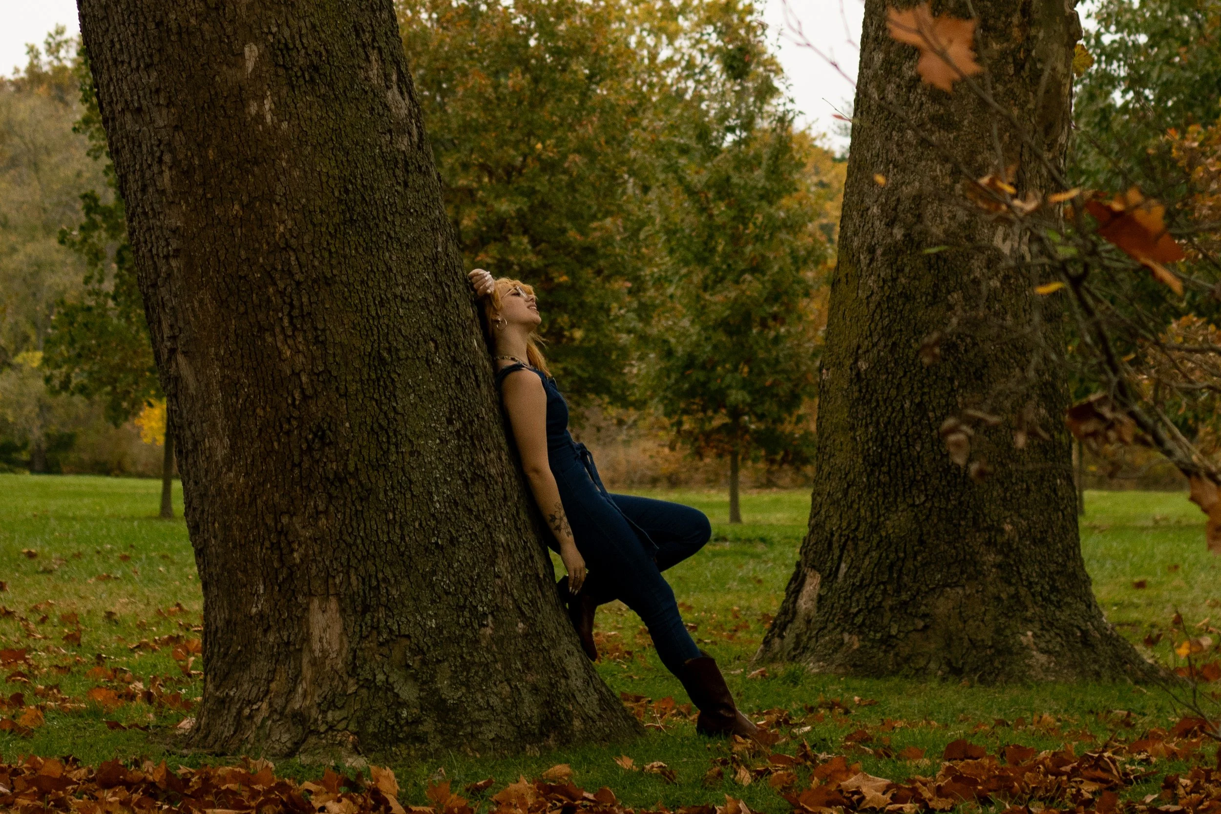 A woman leaning against a tree in a park during autumn, with fallen leaves on the ground and colorful foliage in the background.