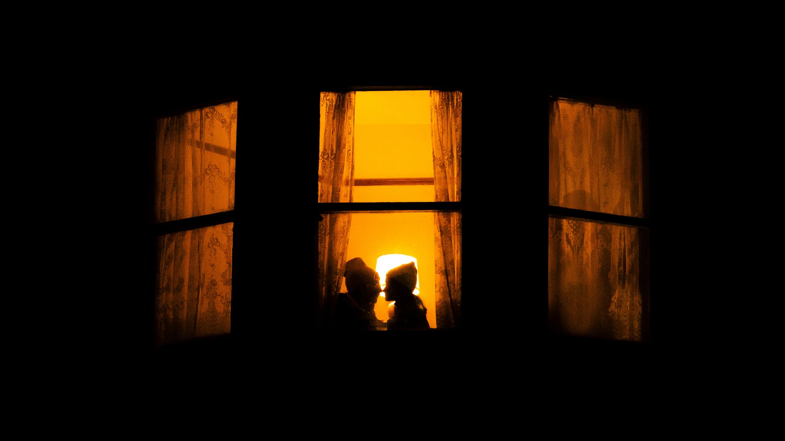 Silhouettes of two people facing each other inside a window with orange curtains, illuminated by warm yellow light from behind.