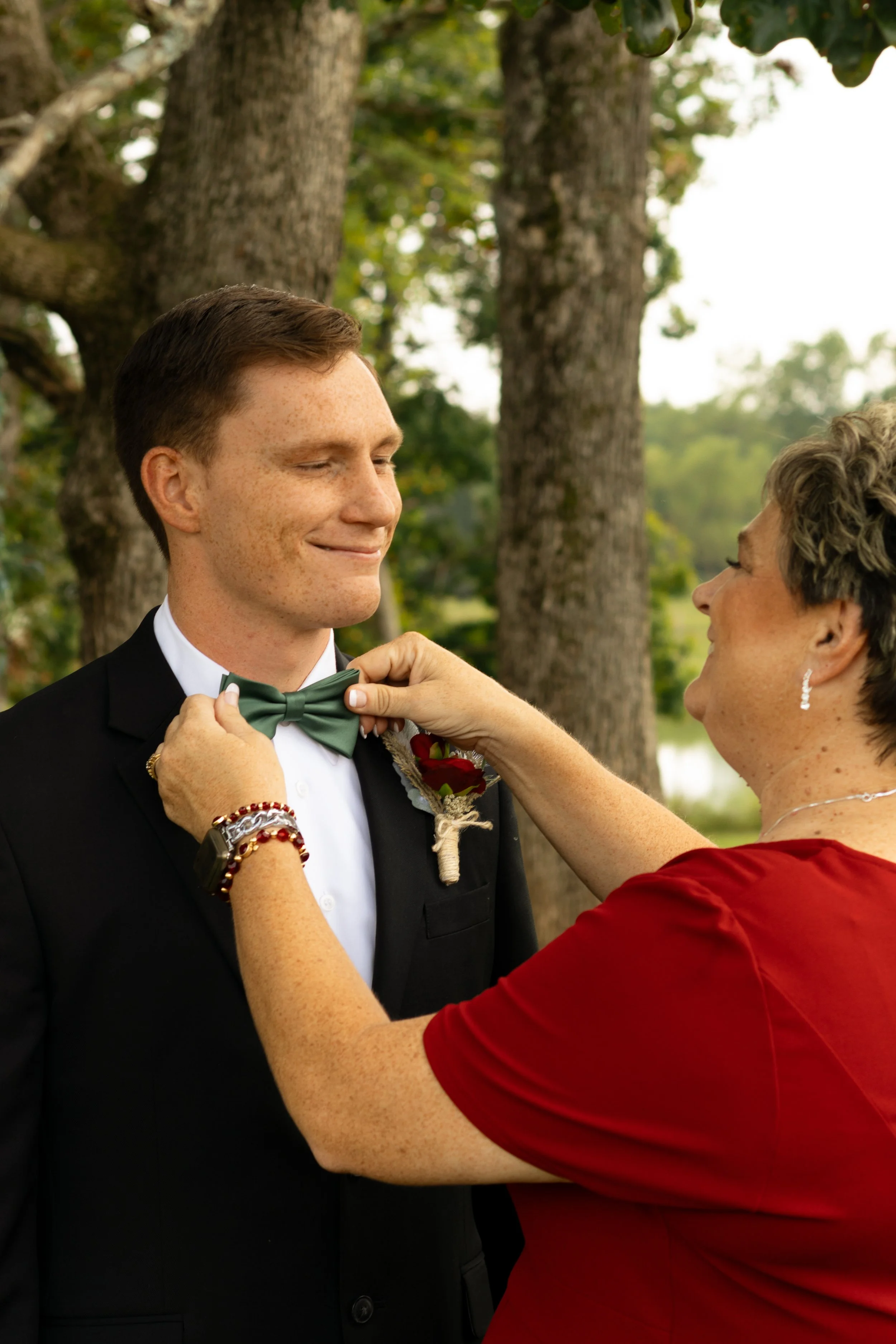 A woman in a red dress adjusting the bow tie of a young man in a black tuxedo during an outdoor ceremony, with trees and a lake in the background.