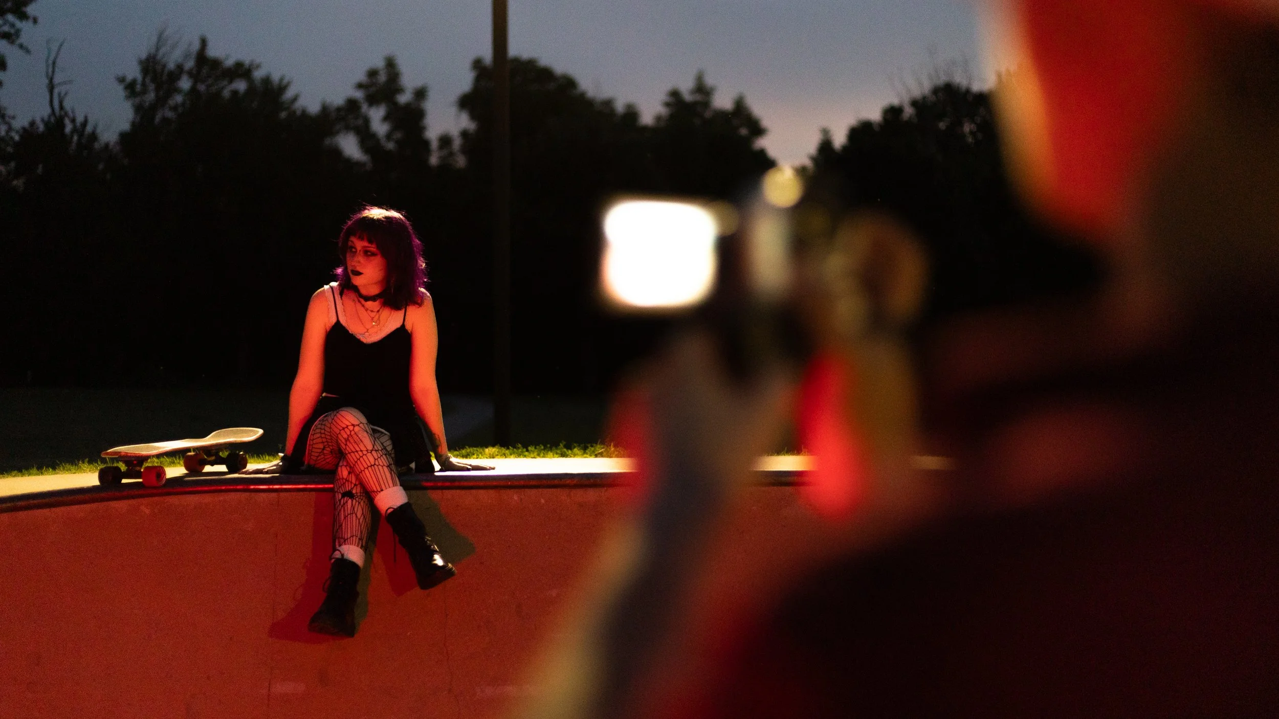 A young woman with black hair, wearing a black dress, fishnet stockings, and combat boots, sitting on the edge of a skateboard ramp during sunset, with a skateboard beside her.