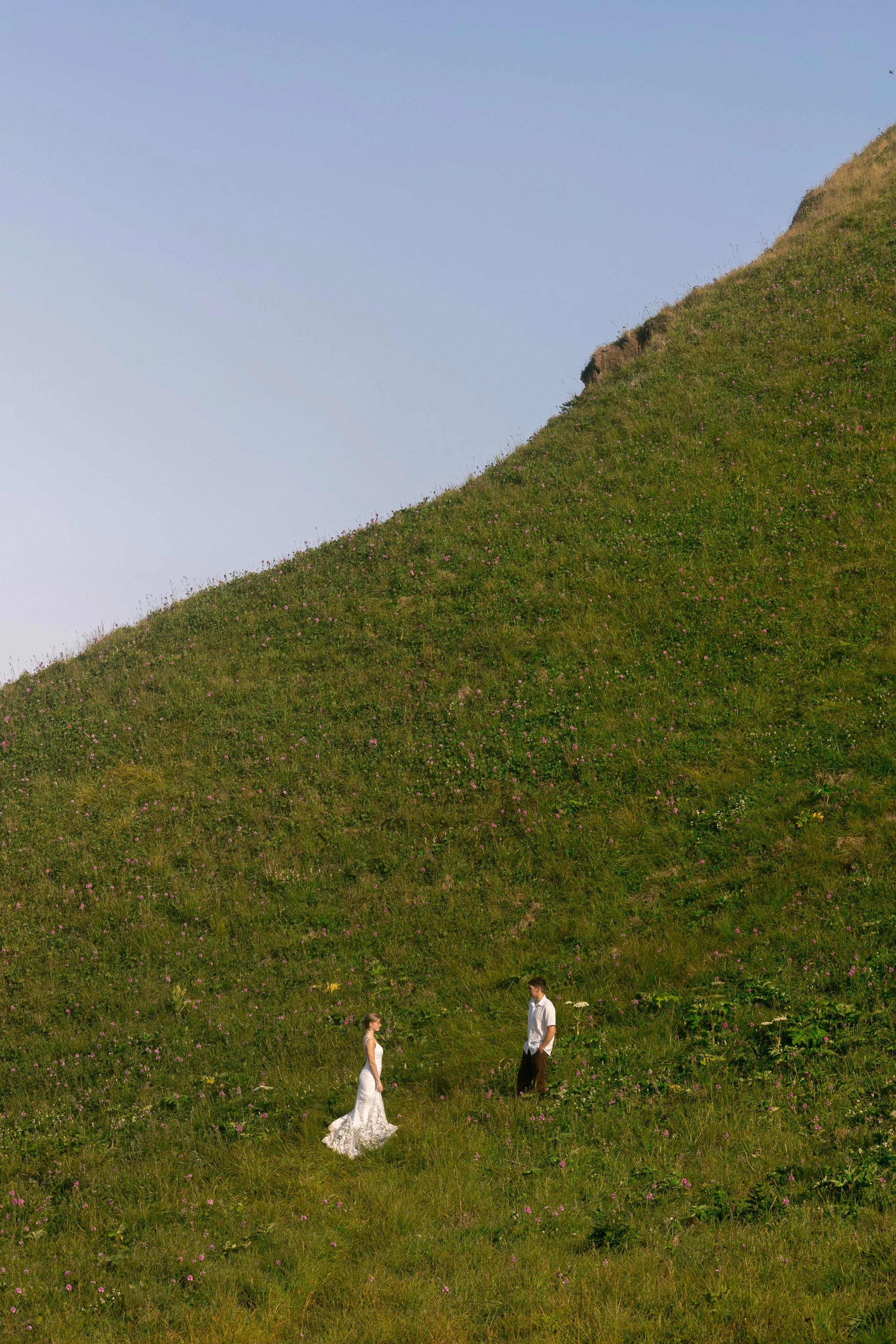 A couple standing on a grassy hillside, with the woman in a white dress and the man in a white shirt and dark pants, against a clear blue sky.