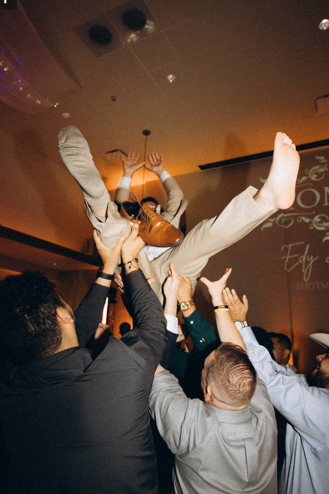 Group of people celebrating at a party, lifting a person in a beige suit with brown shoes into the air.