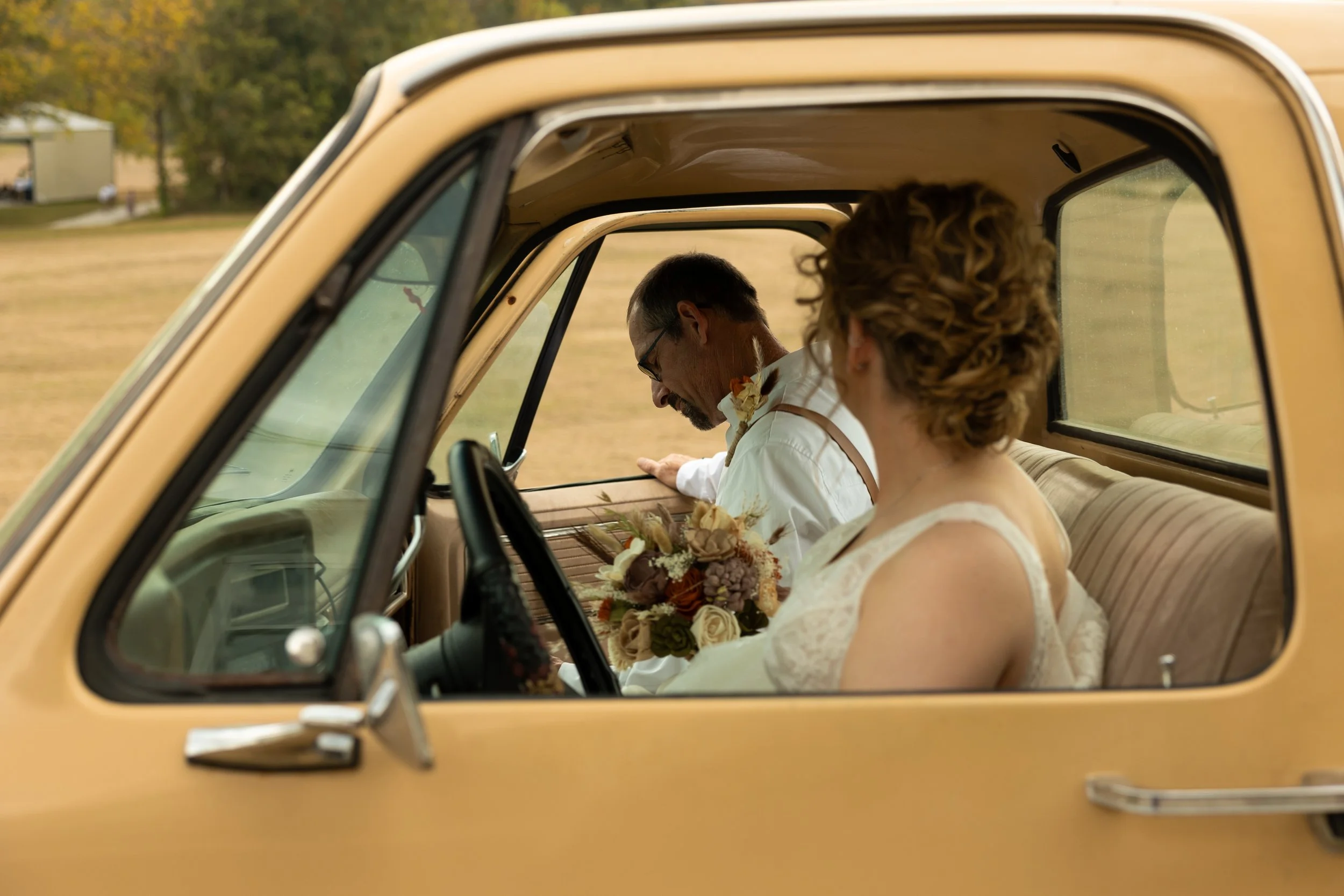 A bride and groom sitting inside a vintage beige car, the bride holding a bouquet of flowers, with a rural landscape in the background.