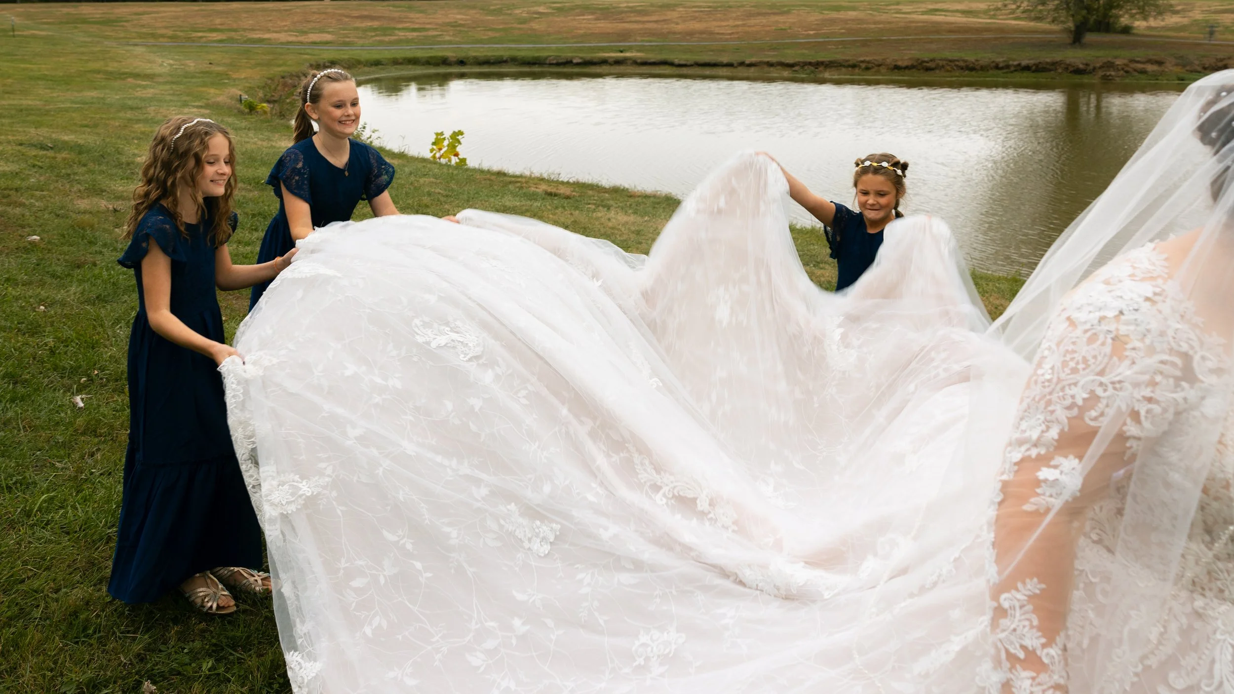 Four young girls in navy blue dresses holding and lifting a white wedding dress near a pond.