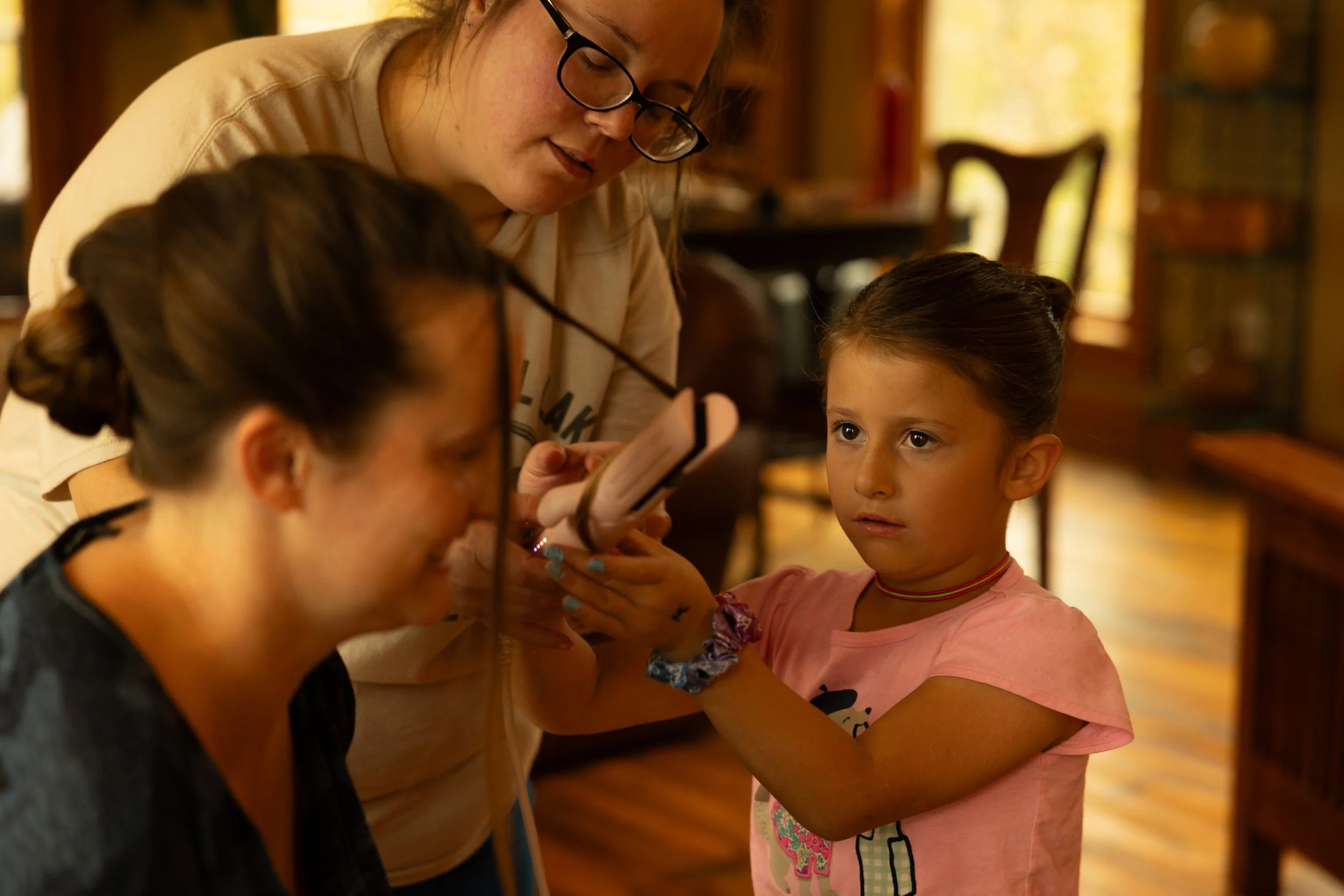 A young girl with a choker necklace and scrunchie on her wrist receives a tattoo from a woman with glasses, while another woman observes, in a cozy room with wooden furniture