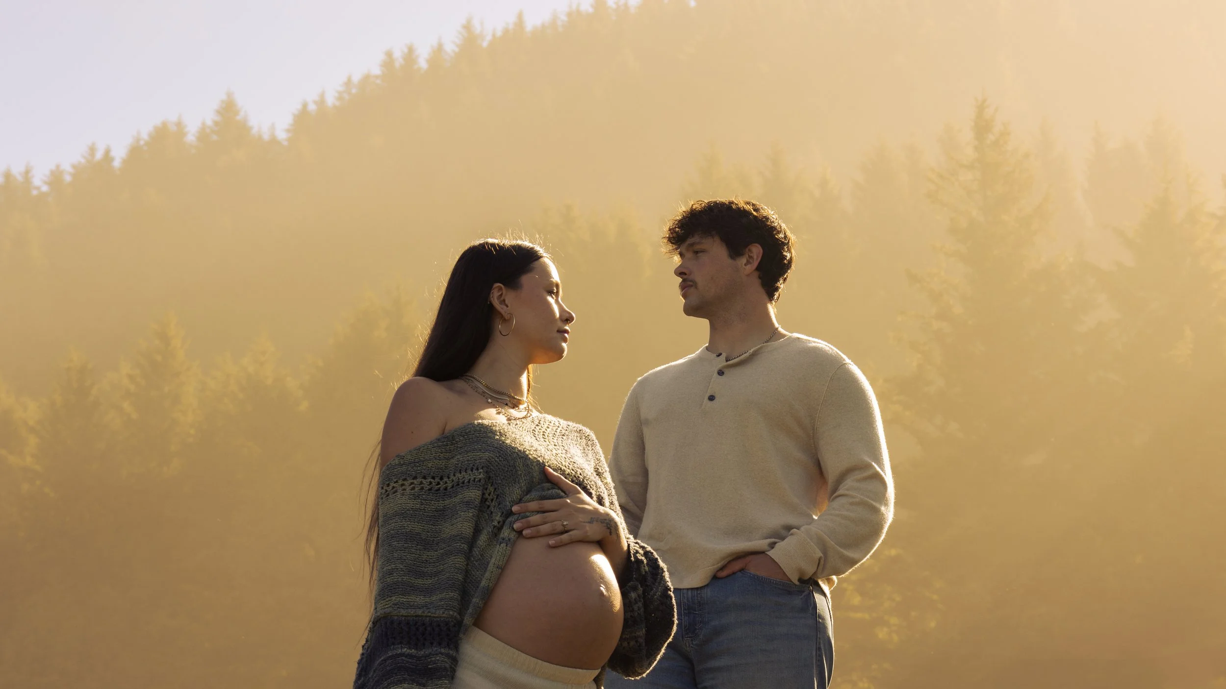 A pregnant woman and a man stand outdoors facing each other during sunset, with a background of trees and a golden sky.