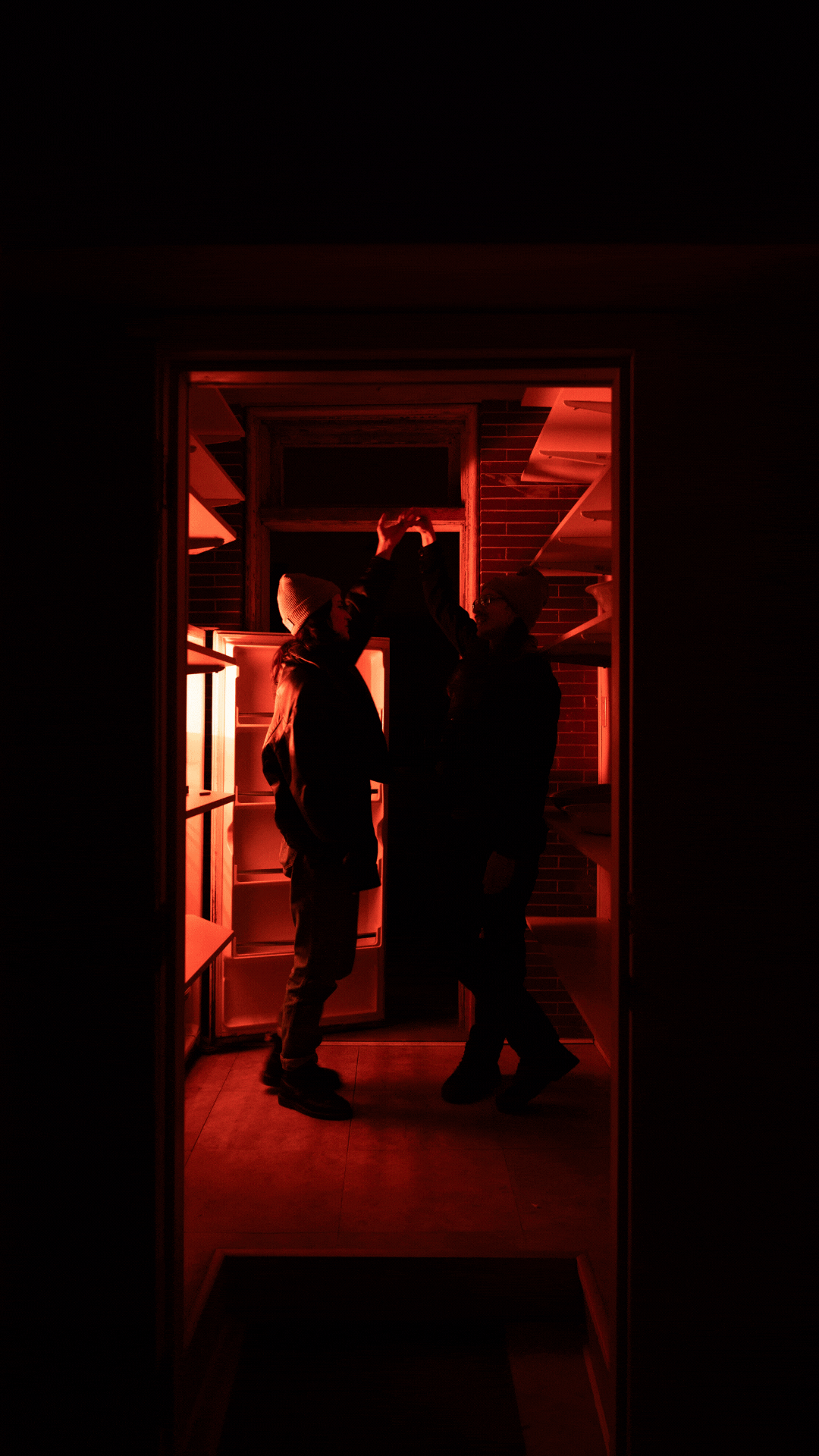 Two people in dark clothing and knit hats dance inside a doorway illuminated by red light, with shelves on either side. One person reaches up while the other smiles at them.