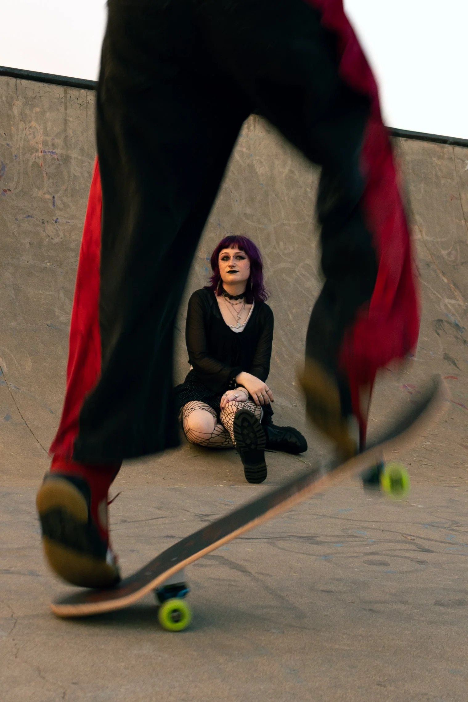 A woman with dark makeup and purple hair sitting on the ground at a skate park, watching a skateboarder with red and black pants skate by.