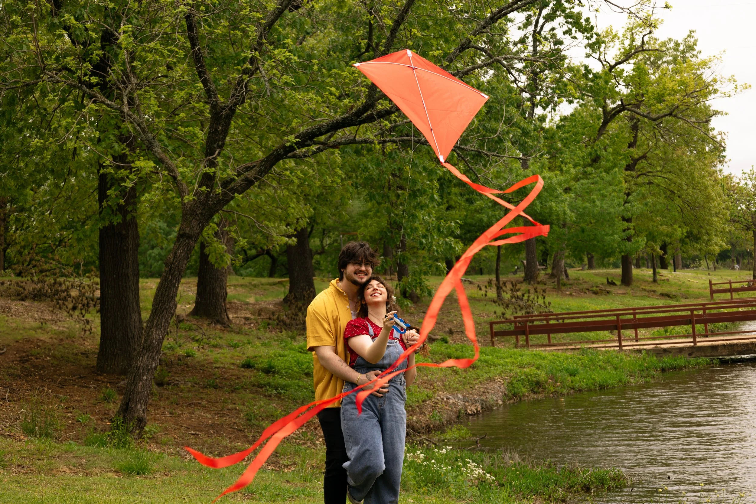 Two people, a man and a woman, are flying a red kite at the park near a pond, surrounded by green trees.