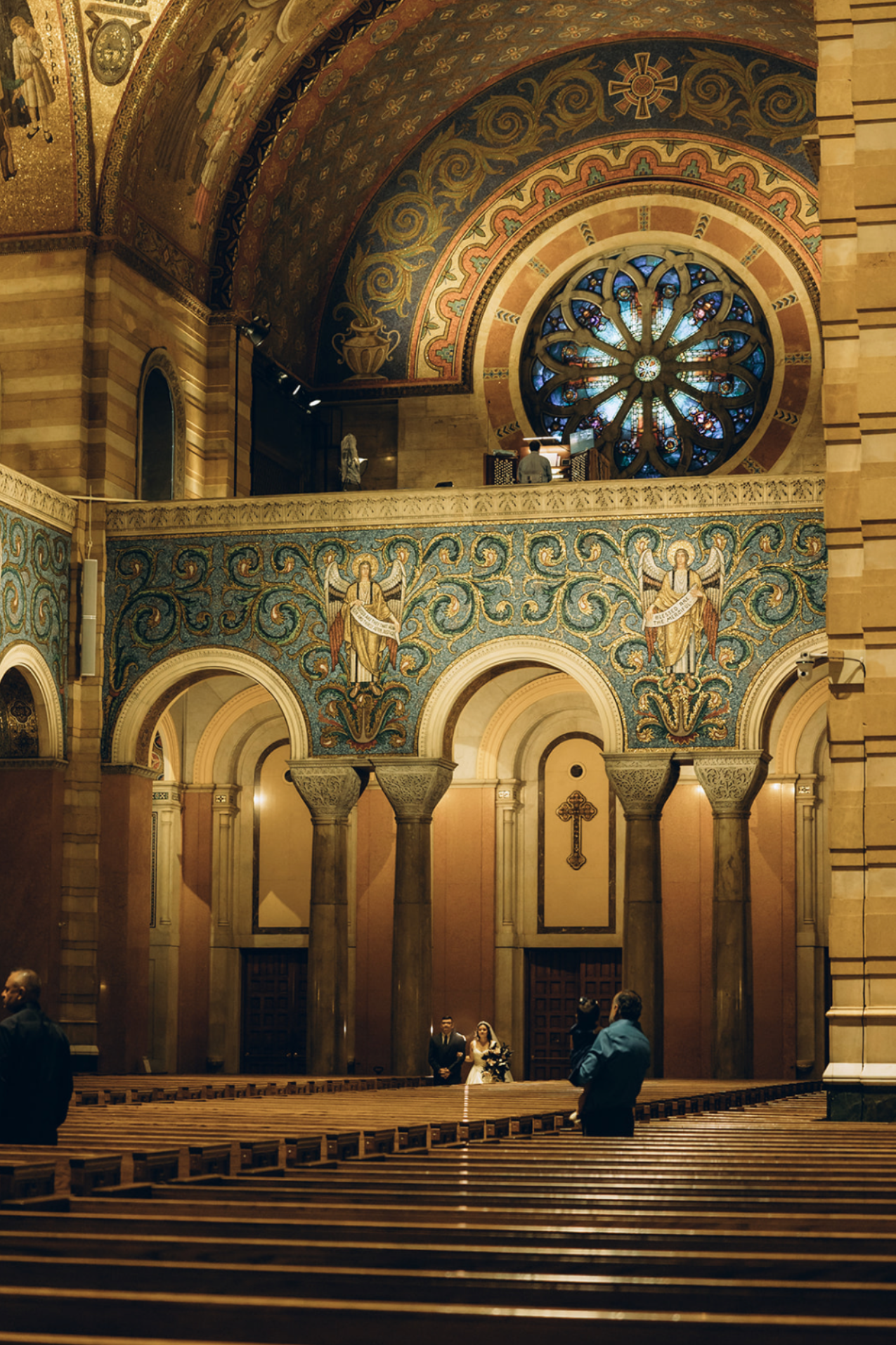 Inside a church with ornate architecture, stained glass window, columns, and a couple standing at the altar.