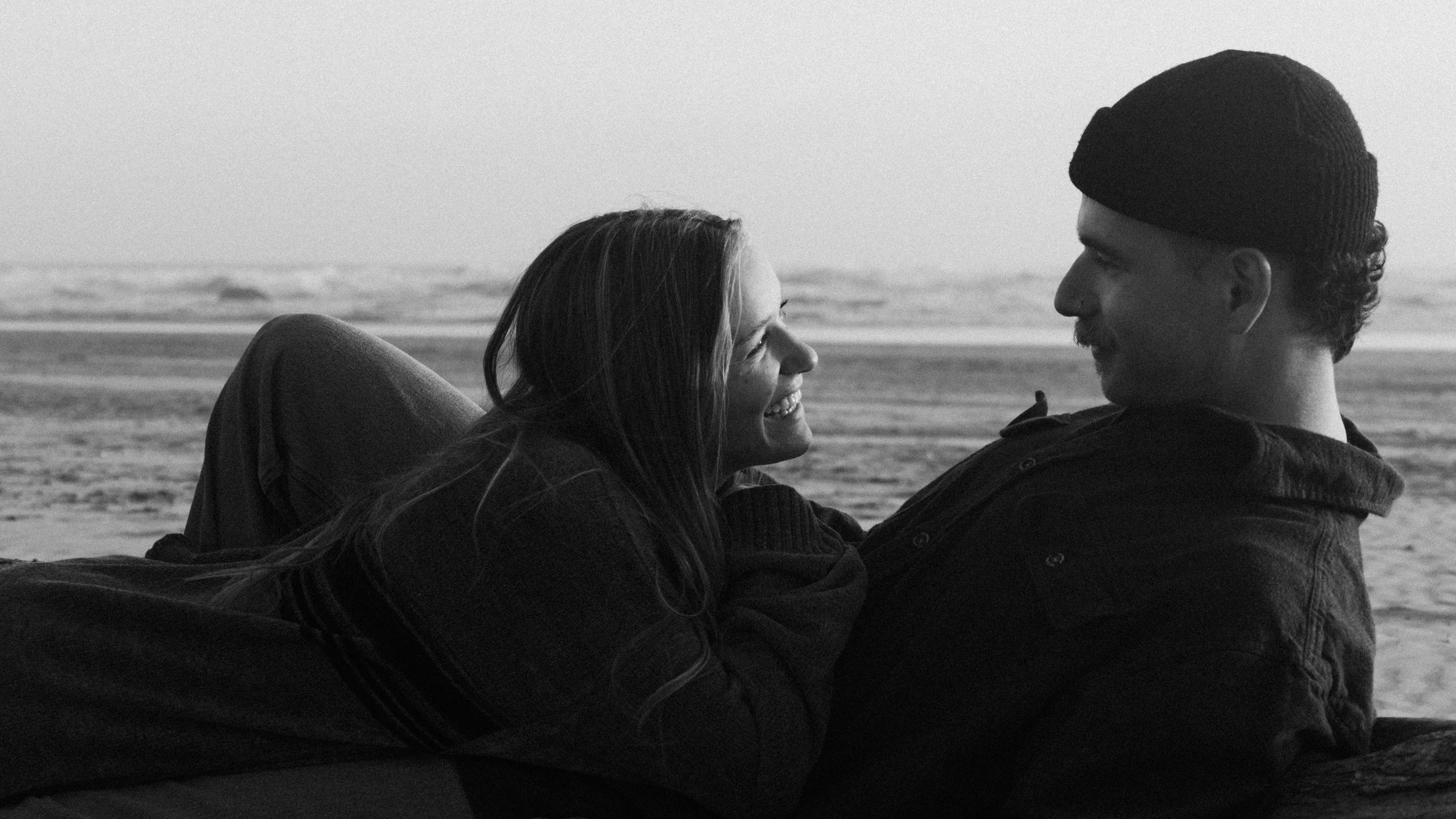 A couple smiling and looking at each other on a beach, lying close to each other with ocean waves in the background.