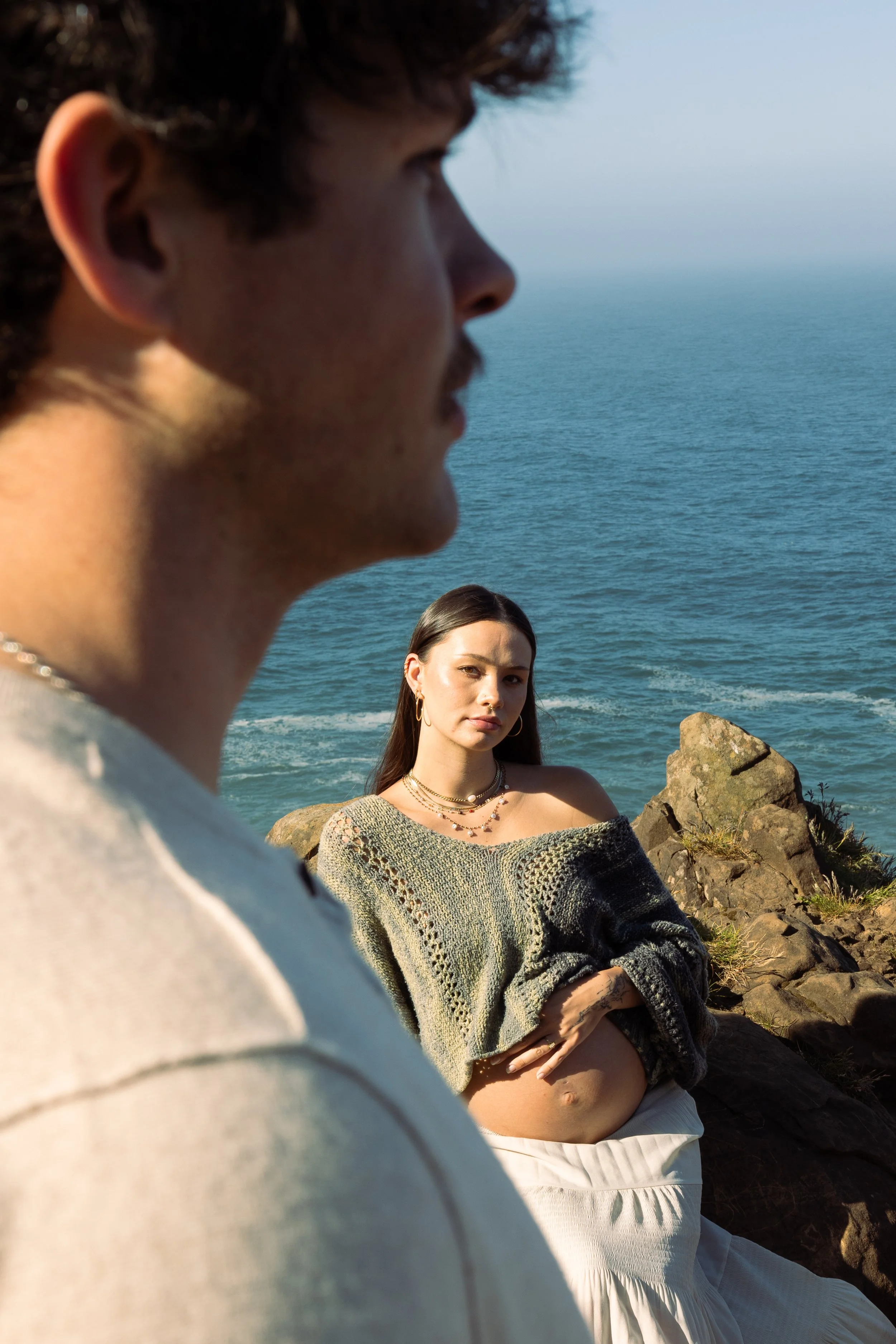 A young man and woman by the ocean, with the man in the foreground out of focus and the woman in the background in focus, sitting on rocks near the water, wearing casual clothes and jewelry.