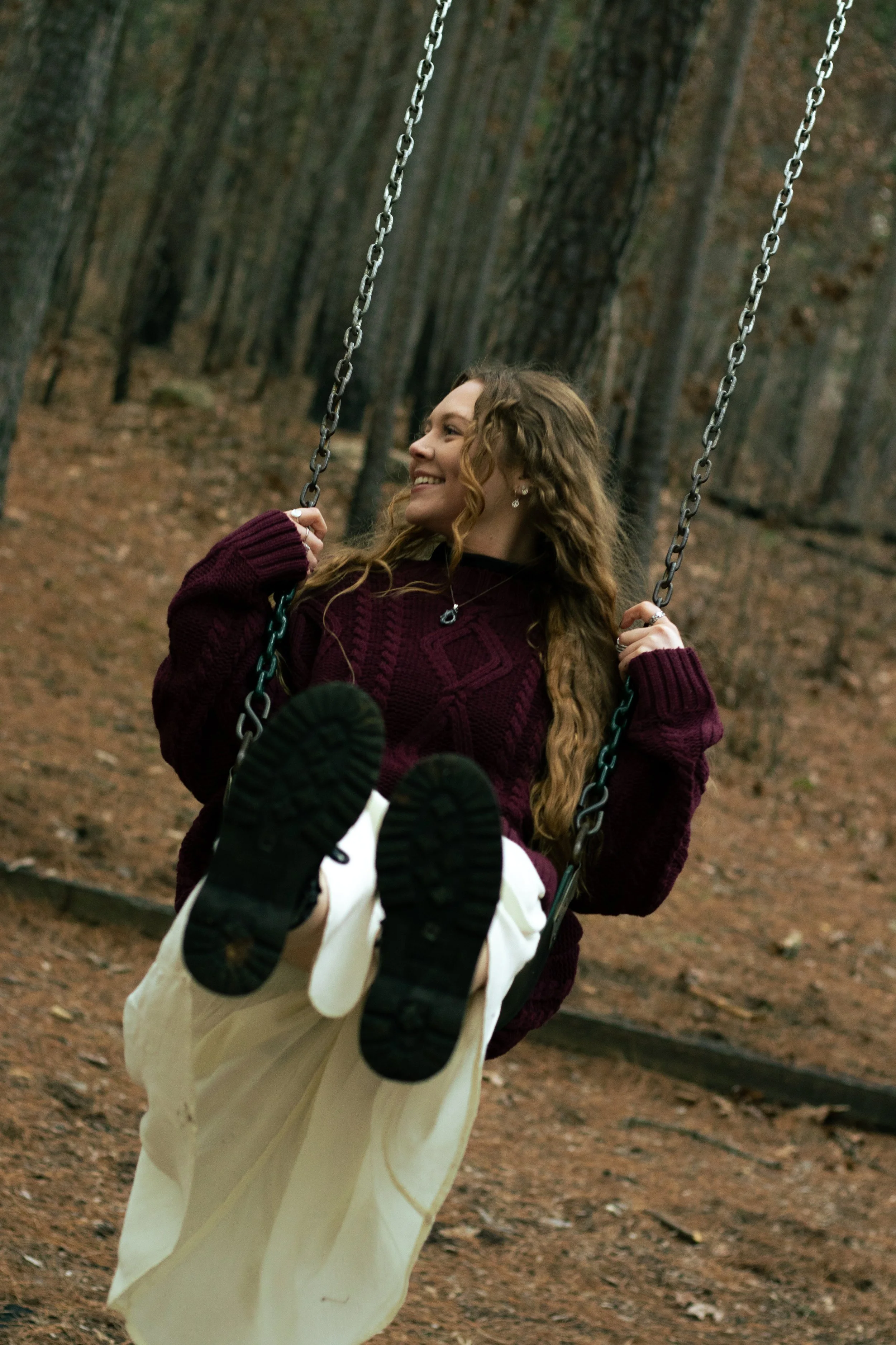 A young woman with long curly hair wearing a maroon sweater and white pants is sitting on a swing in a wooded park, smiling and enjoying the outdoors.