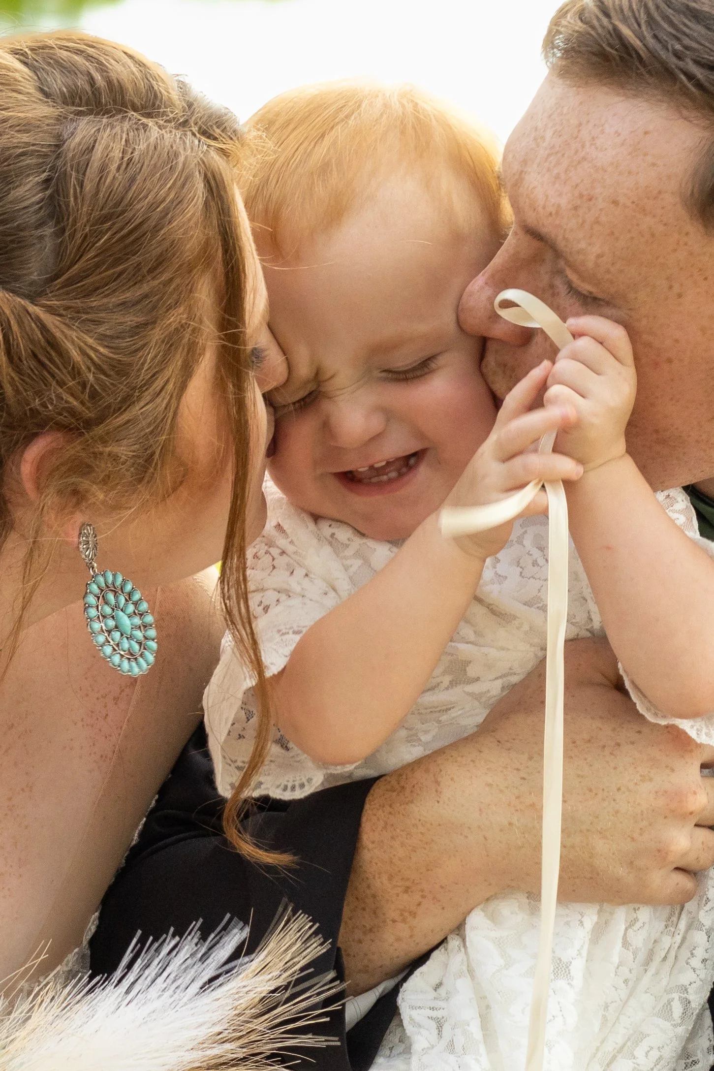 A family embracing each other closely, with a young girl in the middle smiling and laughing, a woman and a man kissing her cheeks in a loving embrace.