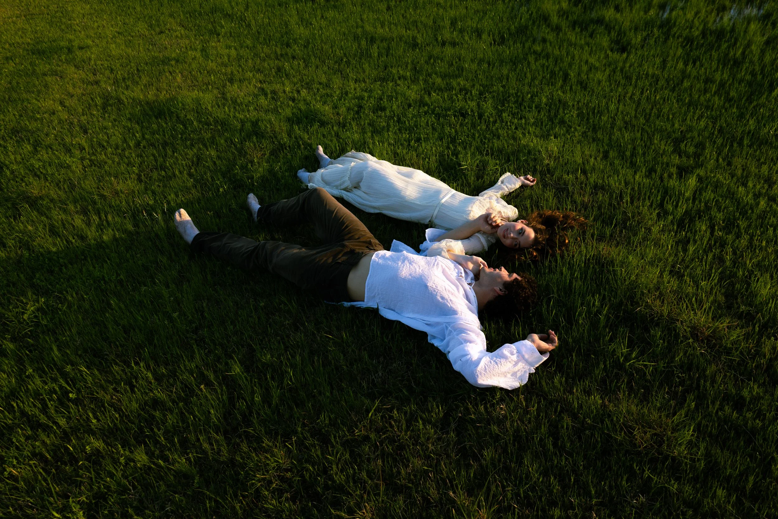 A couple lying on a grassy field during sunlight, with the woman in a white dress and the man in a white shirt and dark pants, holding hands and facing each other.