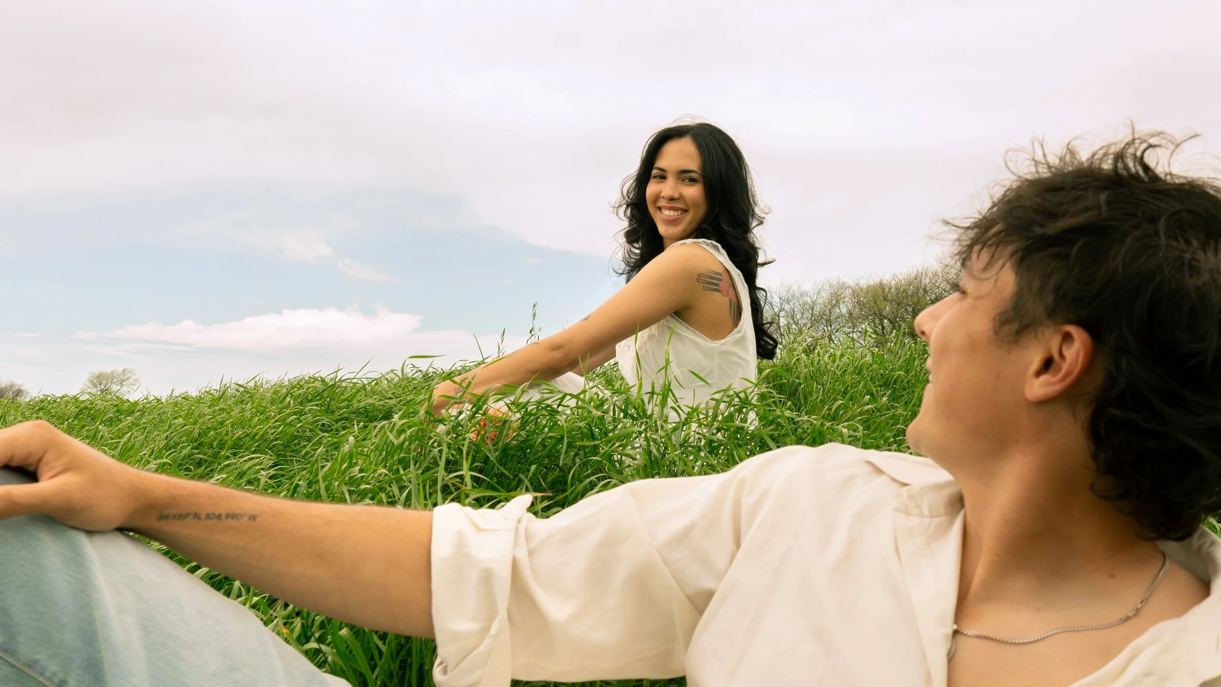 A smiling woman with dark wavy hair sitting in a grassy field and looking at a man lying in the grass, who is also smiling and looking at her.