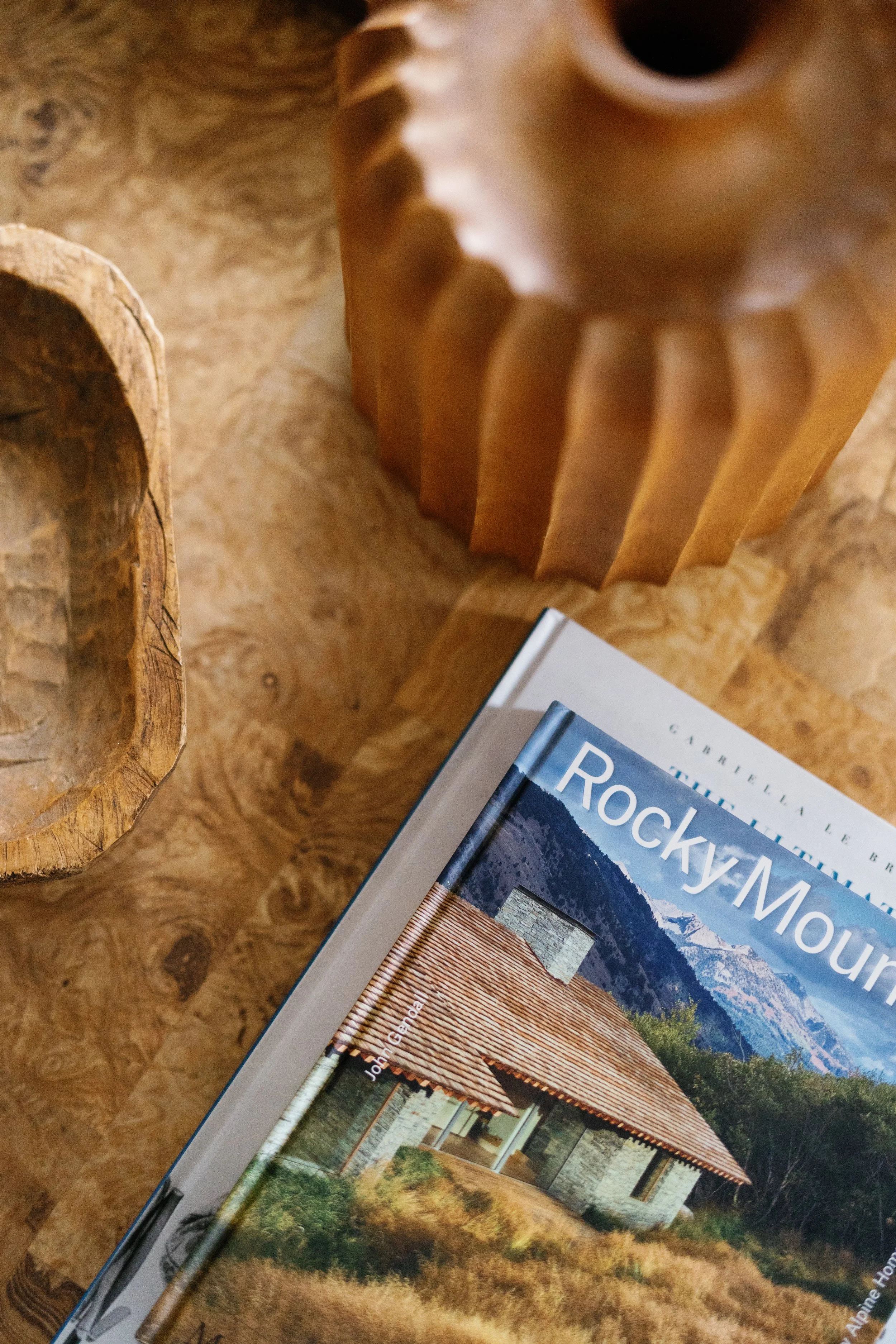 Close-up of a hardcover book titled 'Rocky Mountain' with a photograph of a house and mountains on the cover, placed on a wooden table with carved wooden objects nearby.