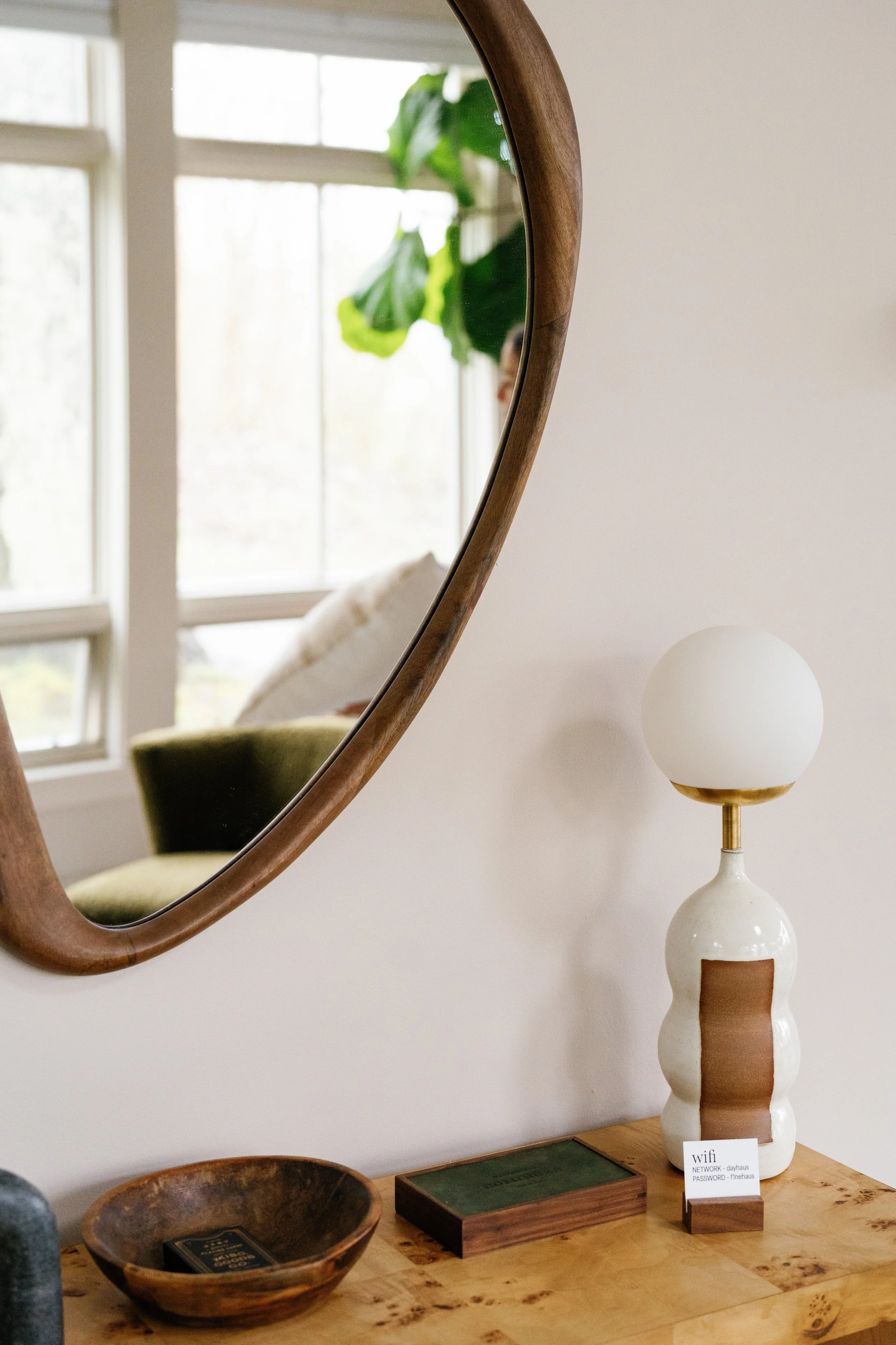 A close-up of a wooden-framed mirror hanging on a white wall, reflecting a window with a green plant and a green upholstered chair. On a wooden surface below the mirror, there is a ceramic table lamp with a round white shade, a dark wooden bowl, a small wooden block with a Wi-Fi sign, and a closed book or box.