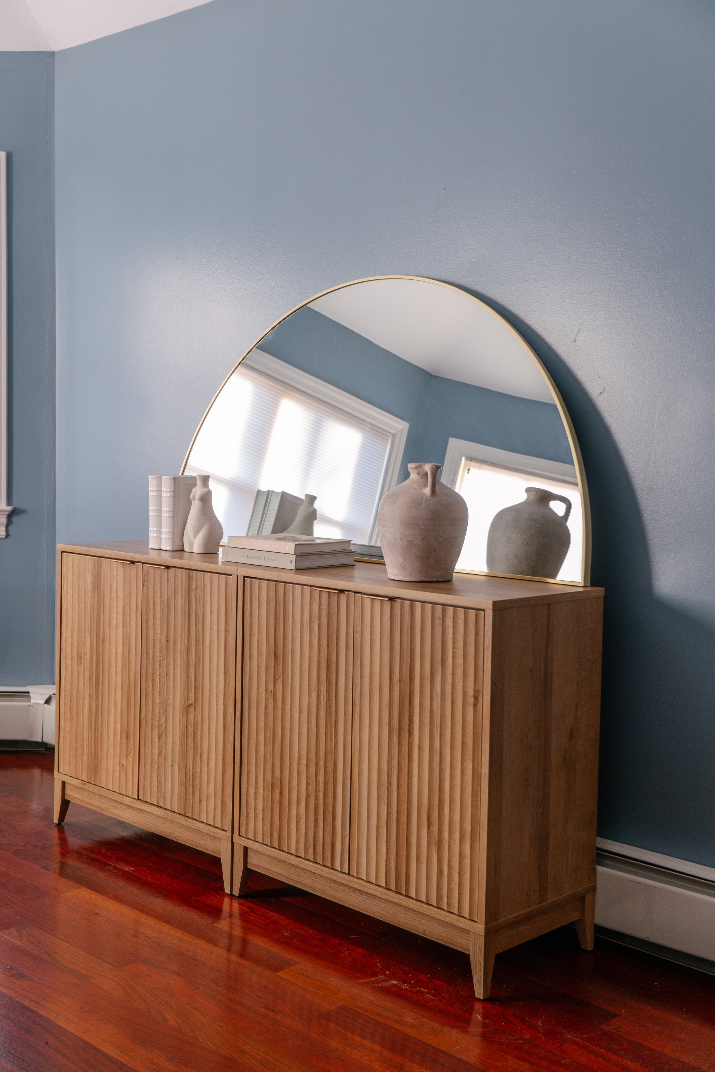 A wooden sideboard with vertical slats and tapered legs, topped with white and gray vases, books, and a large arched mirror, in a room with blue walls and hardwood floors.
