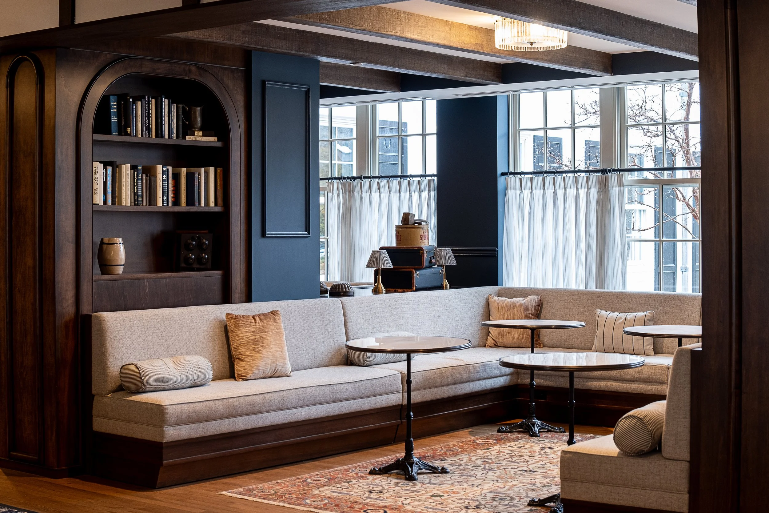 A cozy living room in a hotel lobby with a built-in beige sofa, wooden tables, pillows, built-in bookshelf, blue accent wall, large windows with white curtains, and a patterned rug.