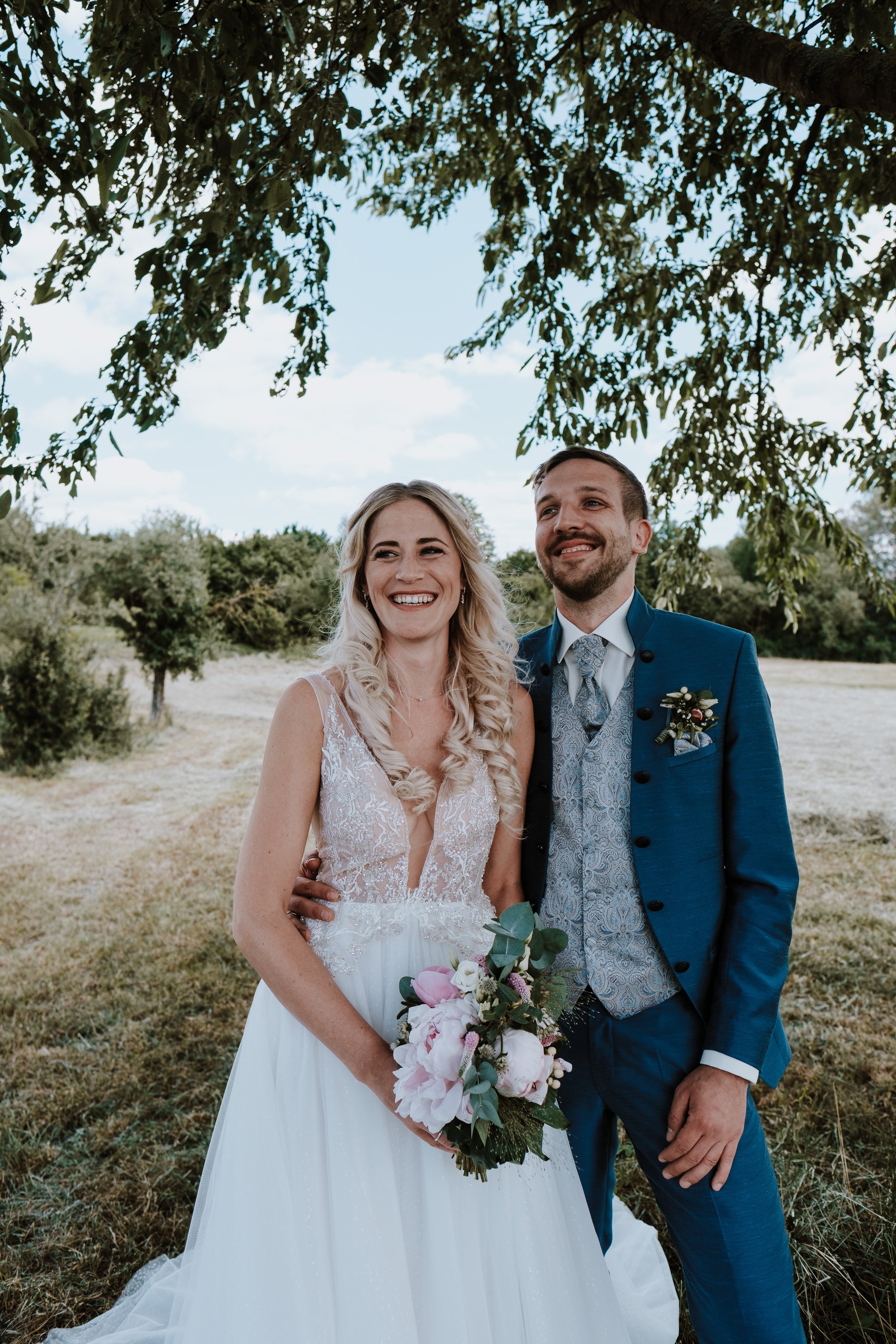 A smiling bride and groom standing under a tree outdoors on their Hamptons wedding. The bride is wearing a white lace wedding dress and holding a bouquet of pink and white flowers. The groom is dressed in a blue suit with a patterned vest and tie.