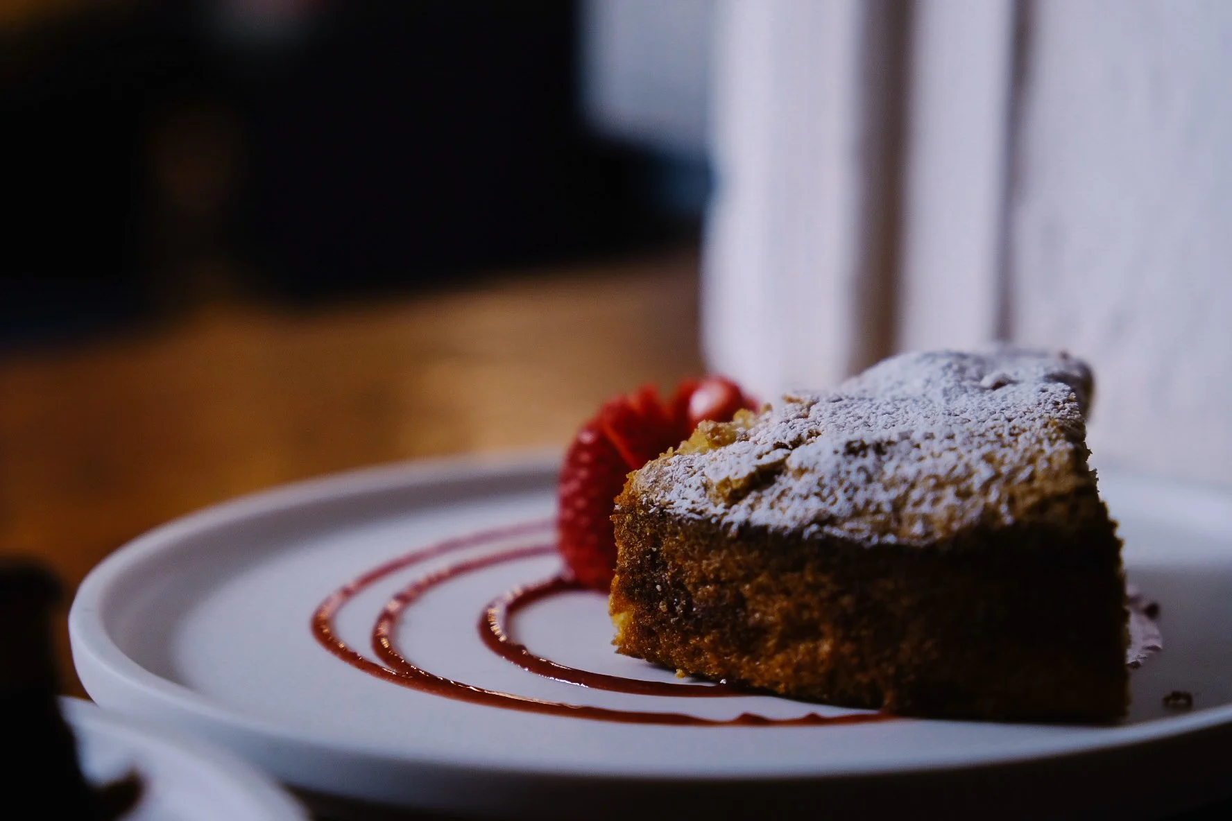 A slice of chocolate cake with powdered sugar topping on a white plate, garnished with a strawberry and drizzled with sauce.