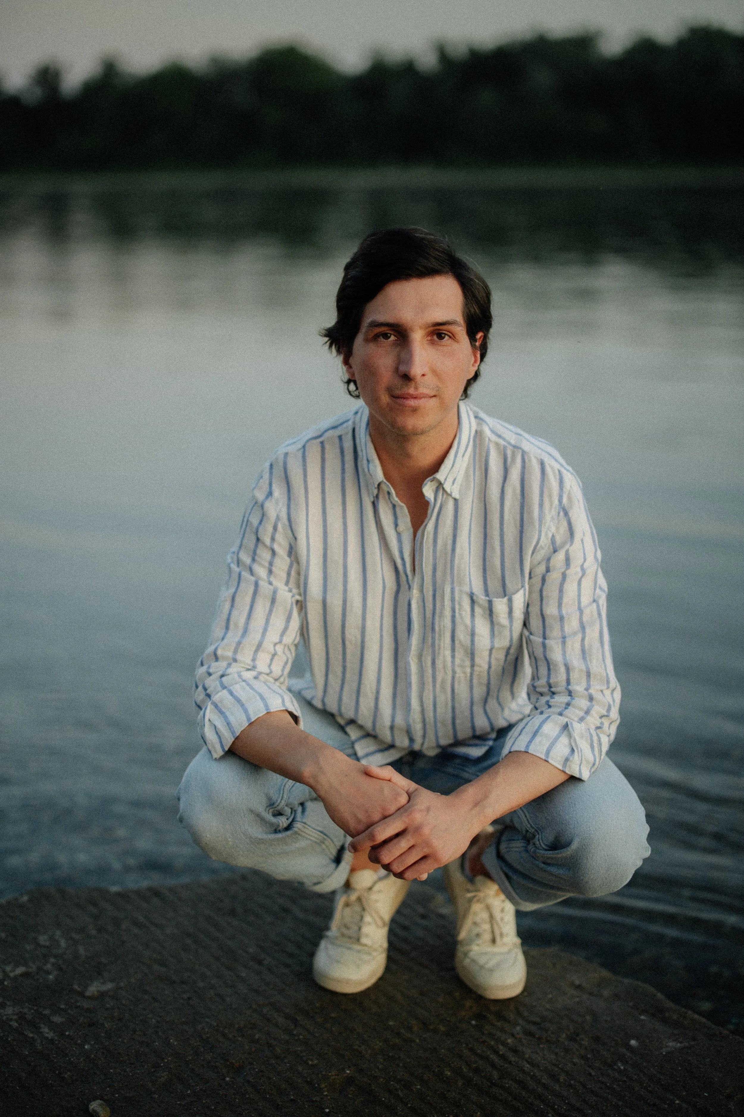 Jair Rojas, the founder of Amauri May Studio in a striped shirt posing on a small dock near a calm body of water during sunset.