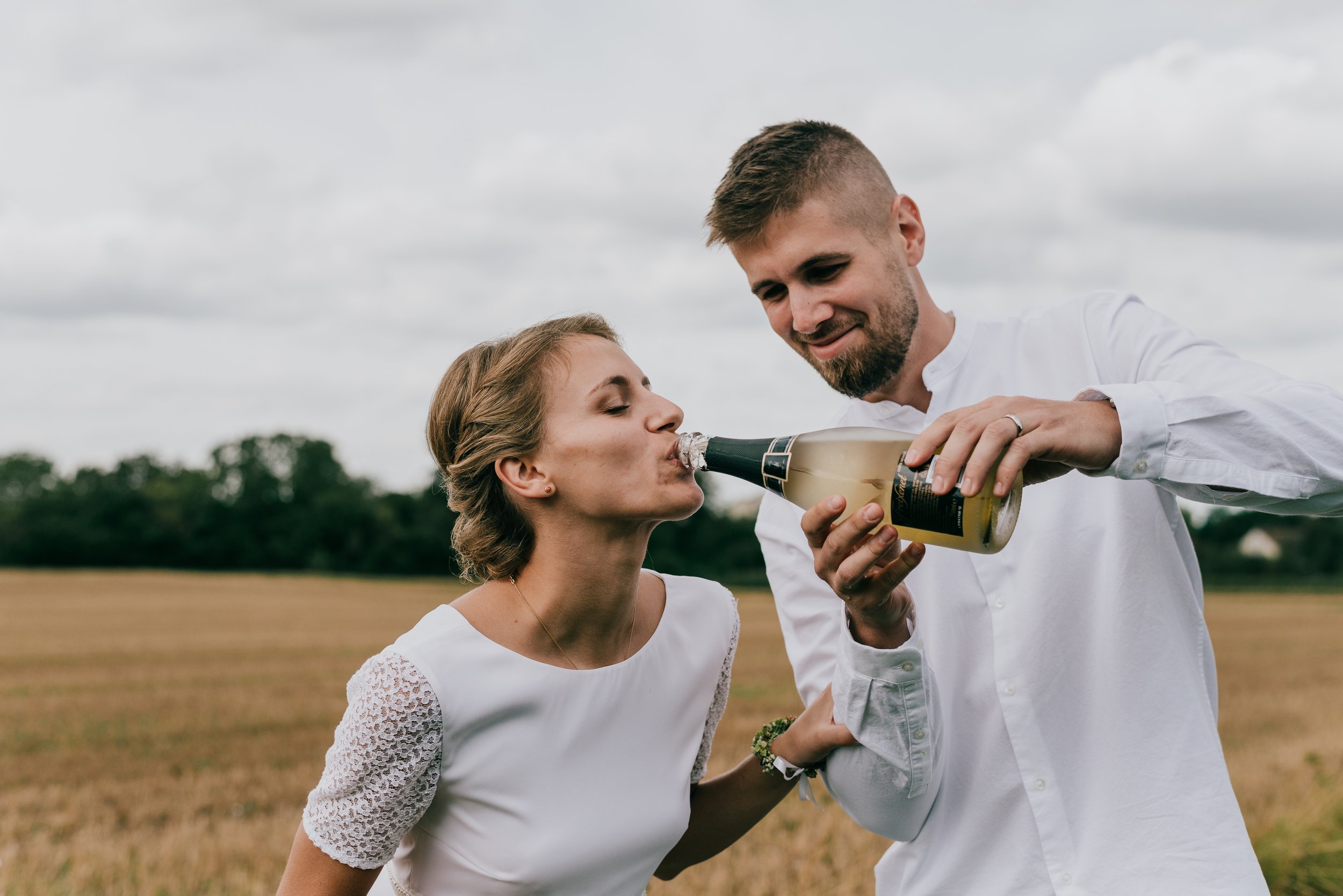 A man and woman dressed in white are outdoors, with the man pouring champagne into the woman's mouth at their destination wedding in Italy. The woman is leaning forward to drink.